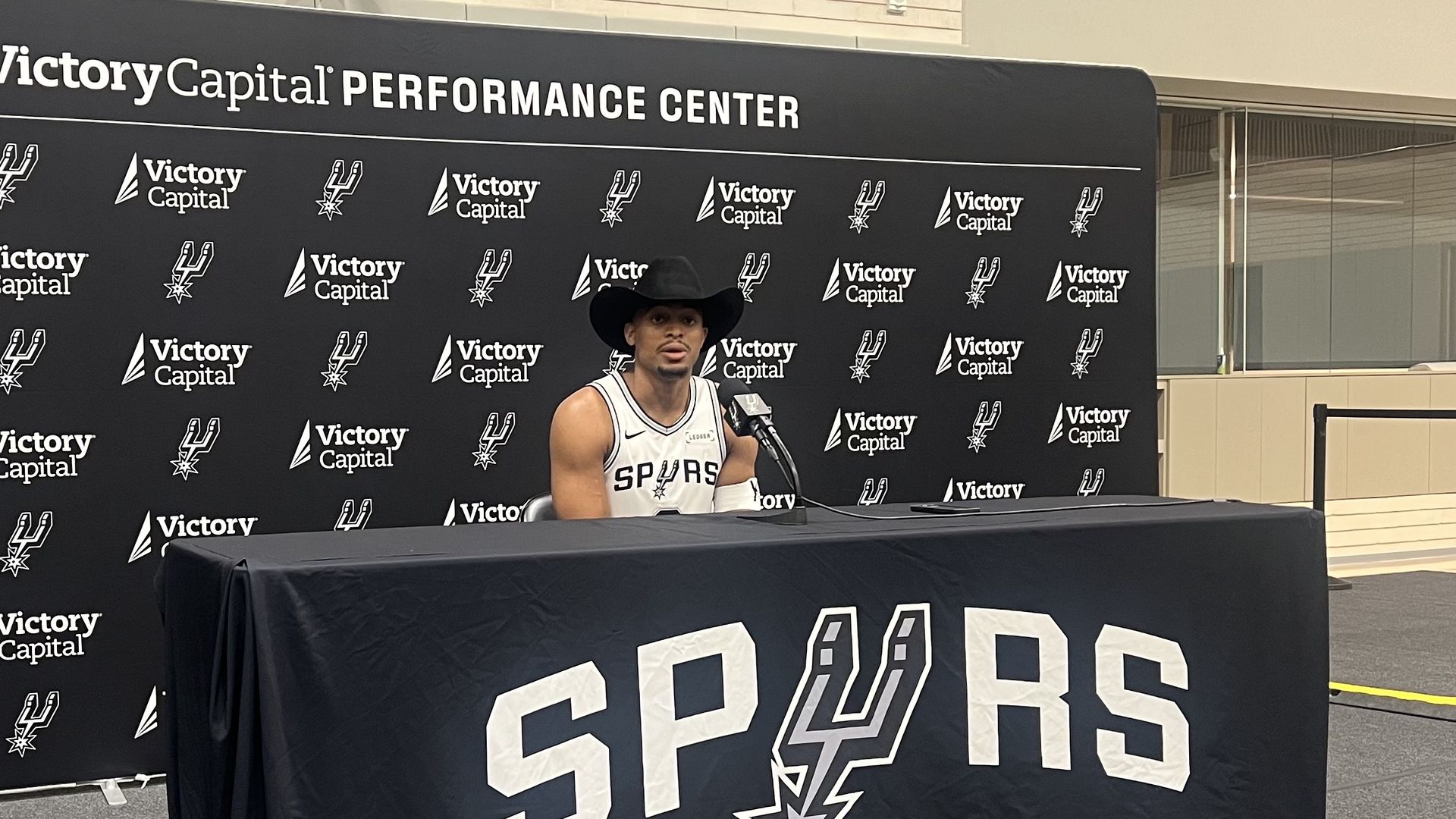 Keldon Johnson, in a white Spurs jersey and black cowboy hat, speaks at a press conference at the Victory Capital Performance Center with Spurs logo backdrop and table cover.