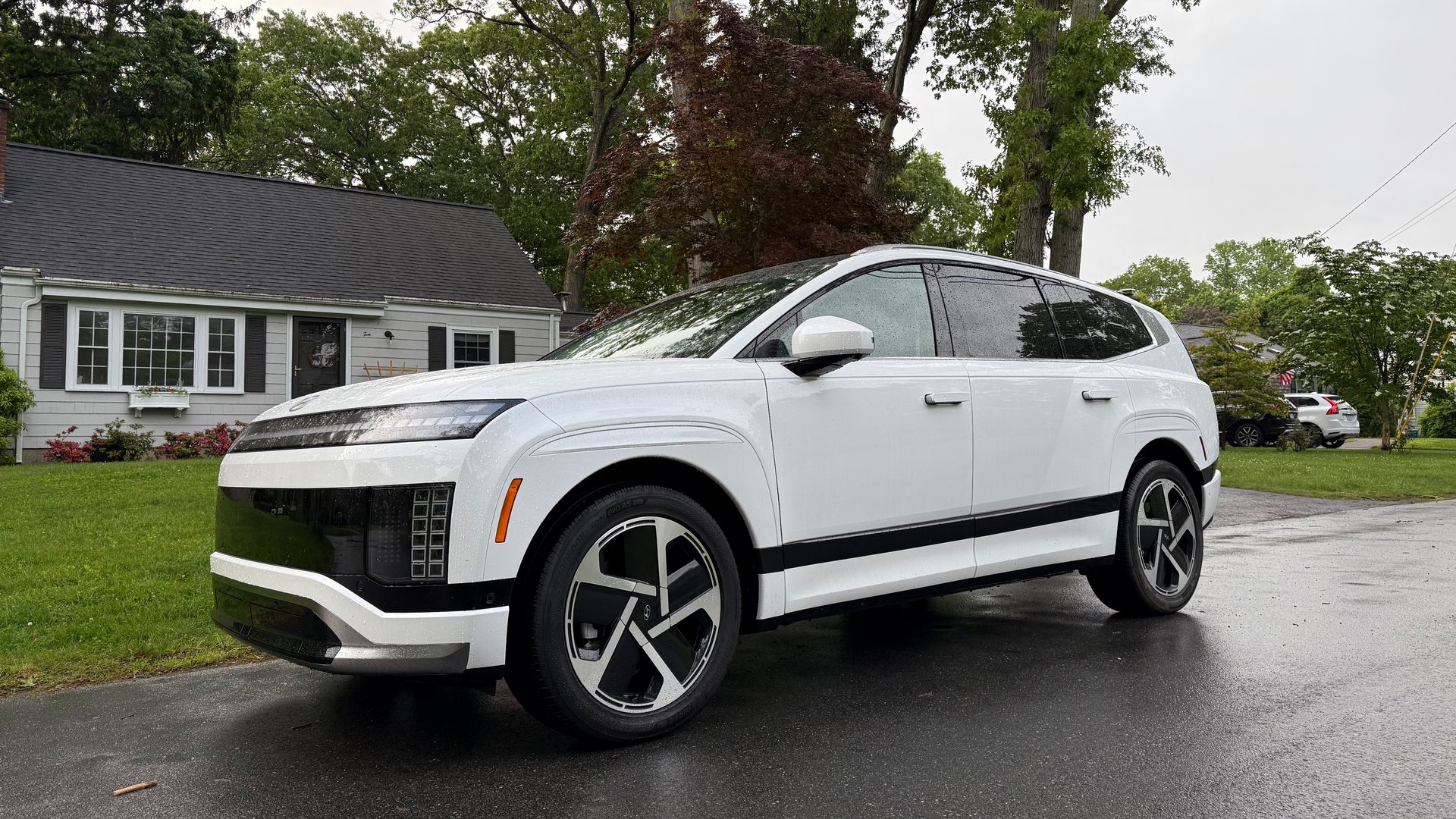 White Hyundai Ioniq 9 electric SUV parked on a wet driveway in front of a suburban house with green lawn and tall trees on a cloudy day.