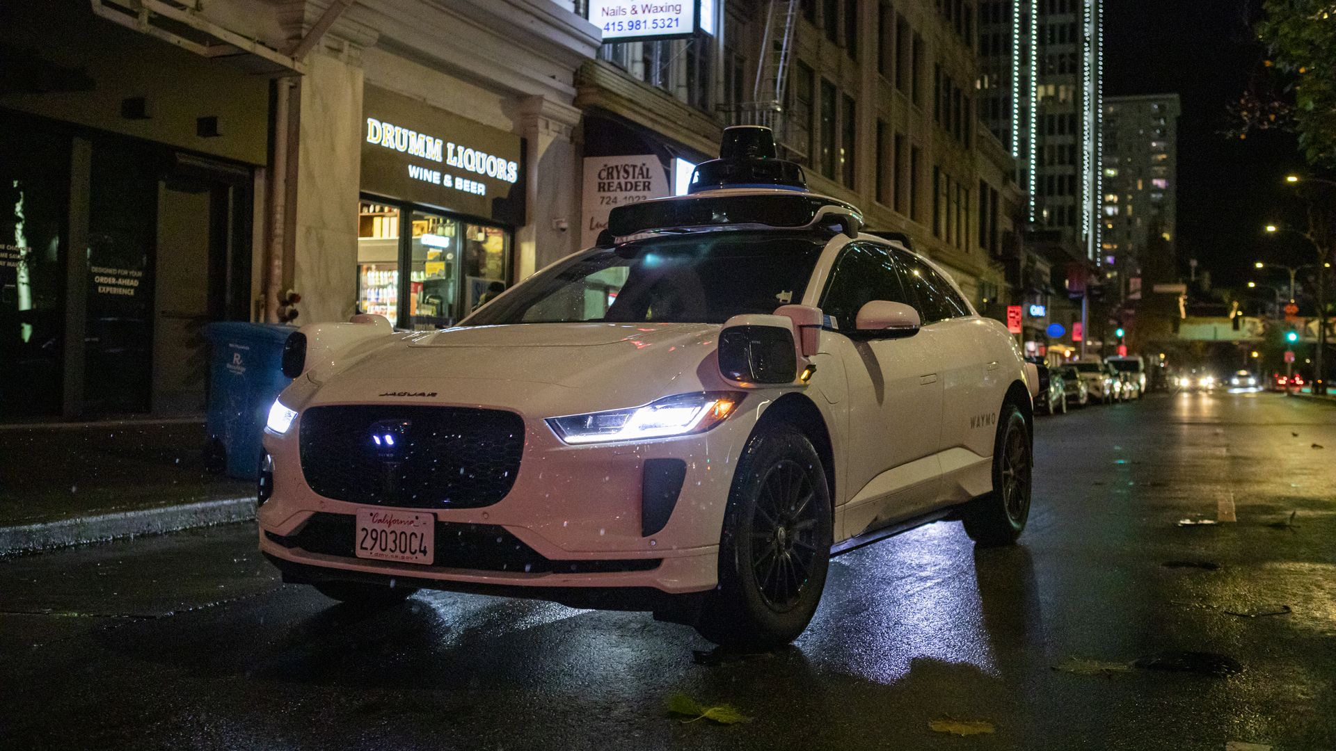 White autonomous Jaguar car with rooftop sensors parked on a wet city street at night, near illuminated shops including "DRUMM LIQUORS" and "Lucky Nails."