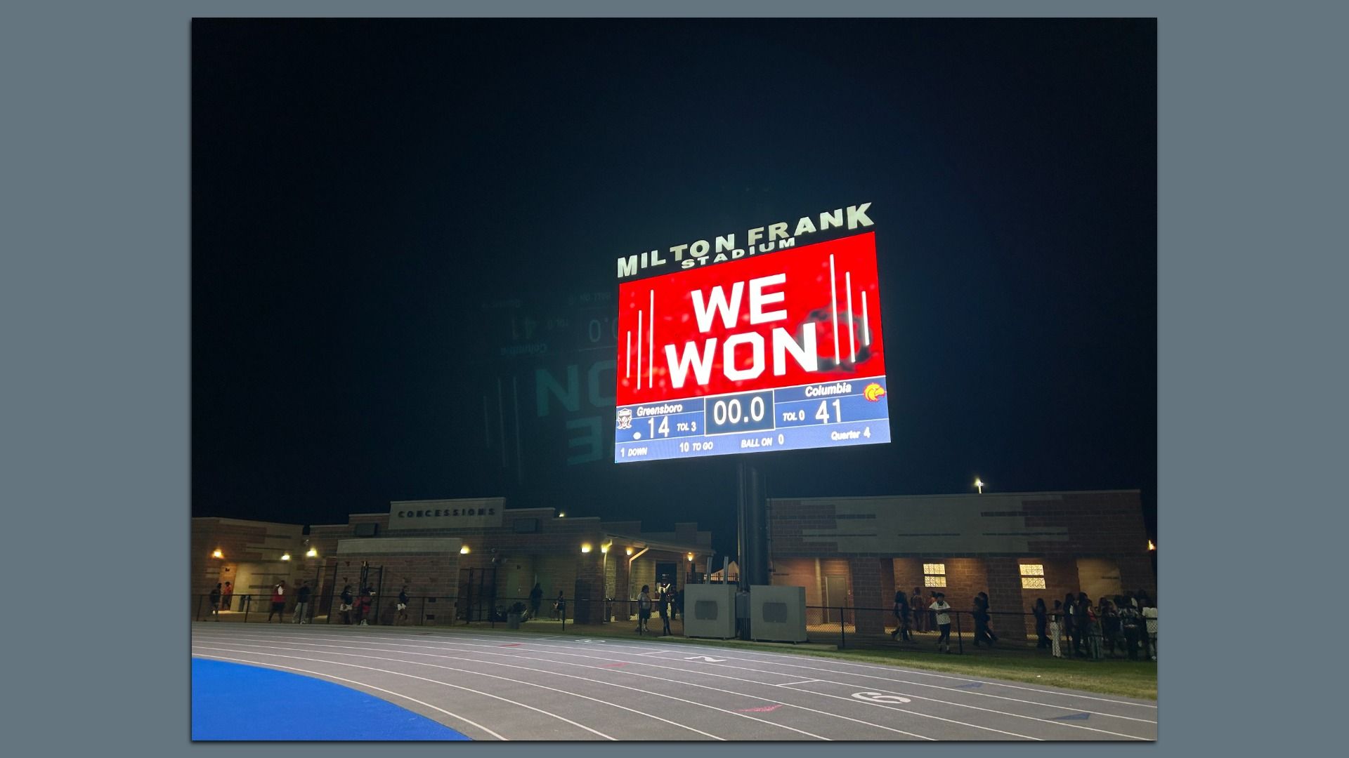 Nighttime view of a stadium scoreboard reading WE WON in white on red; above it reads MILTON FRANK STADIUM. A blue track and brick buildings line the foreground with people.