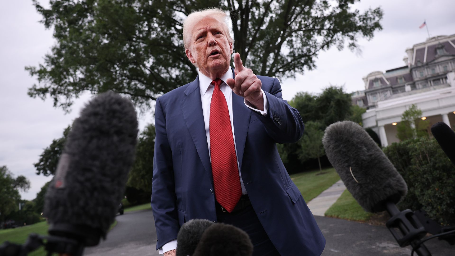 President Trump, dressed in a blue suit, white shirt and red tie, speaking to reporters outdoors, pointing forward, with microphones in the foreground and trees and a building in the background.