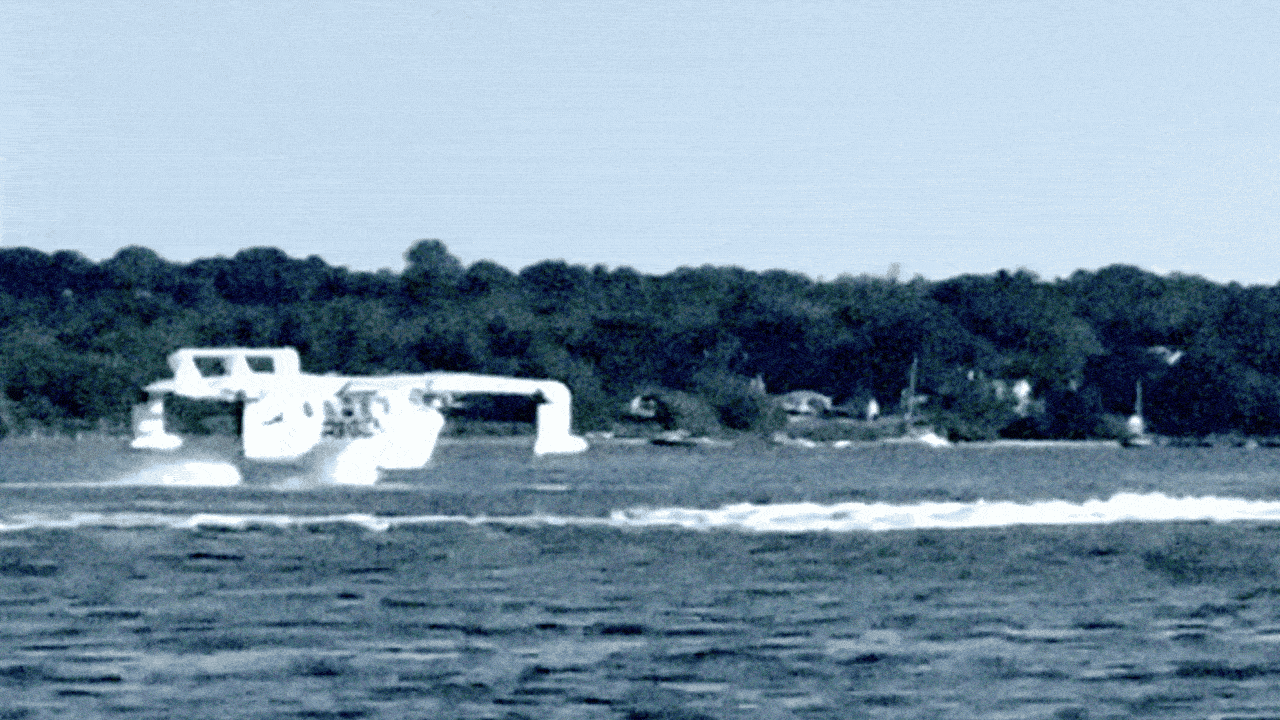 A white boat speeding across blue water with trees and houses visible on the distant shore under a clear sky.