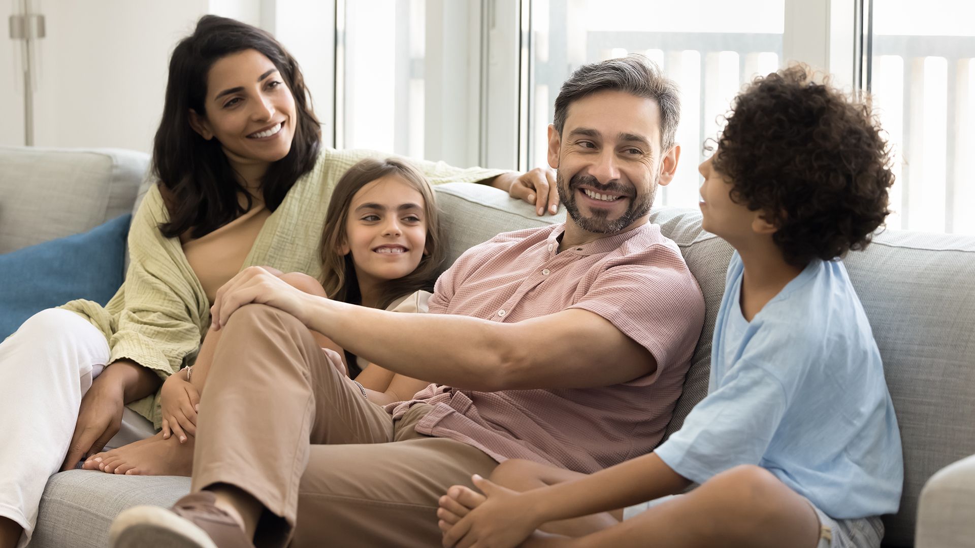 Family of four relaxing on a light gray sofa in a bright living room: mom with dark hair, dad with gray hair and beard in a pink shirt, smiling daughter, and curly-haired son.