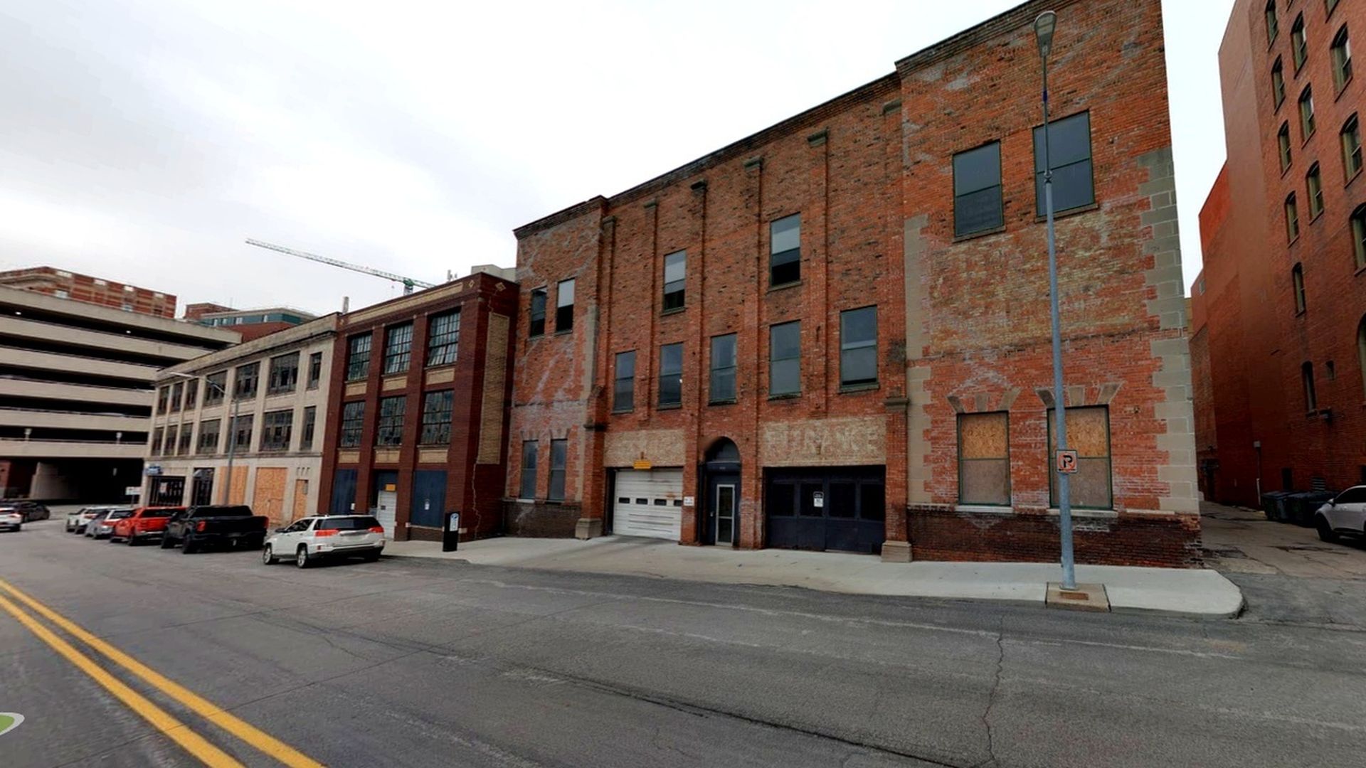 Urban street scene with brick storefronts and a large brick building. Parked cars line the street while a gray parking garage sits to the left; a streetlamp and no-parking sign stand under a cloudy sky.