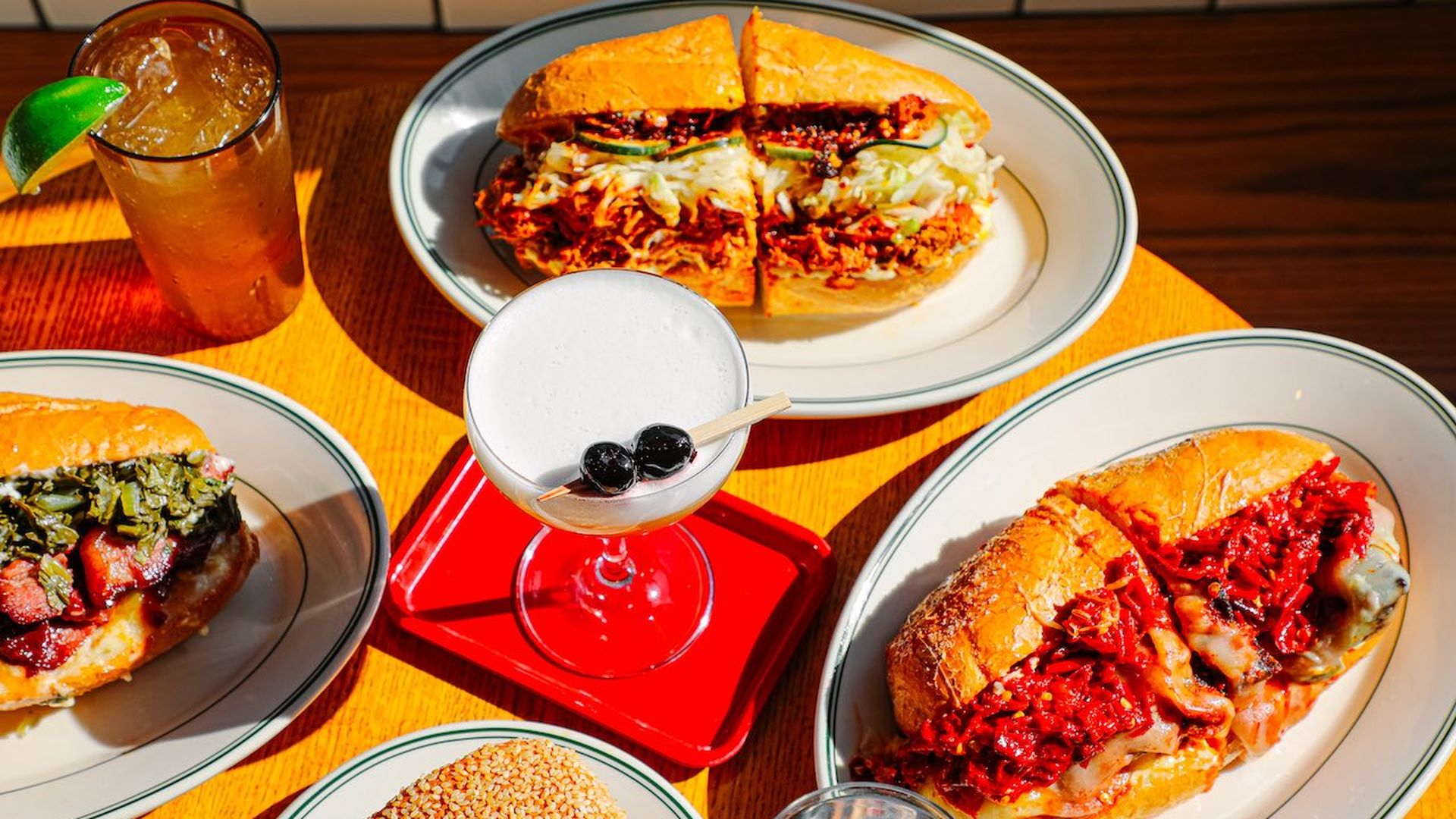 Table with four sandwiches on white plates, including a sandwich with greens and bacon, two sandwiches with melted cheese and red sauce, and a sesame seed bun sandwich. Two drinks, one iced with lime, one white cocktail with two cherries.
