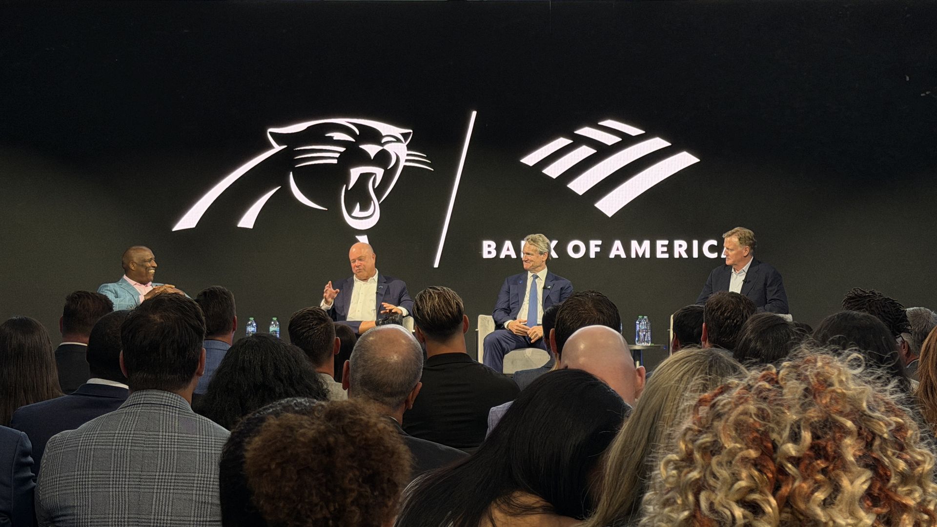 Panel discussion on stage with five men in suits against a Bank of America backdrop featuring a snarling panther logo; audience in foreground.