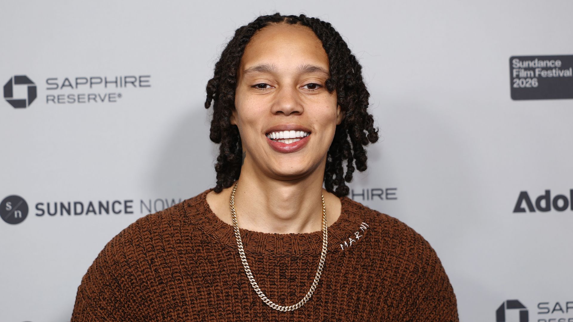 Person with curly hair wearing a brown knit sweater and gold chain necklace smiling in front of a backdrop with Sundance Film Festival 2026 and sponsor logos.