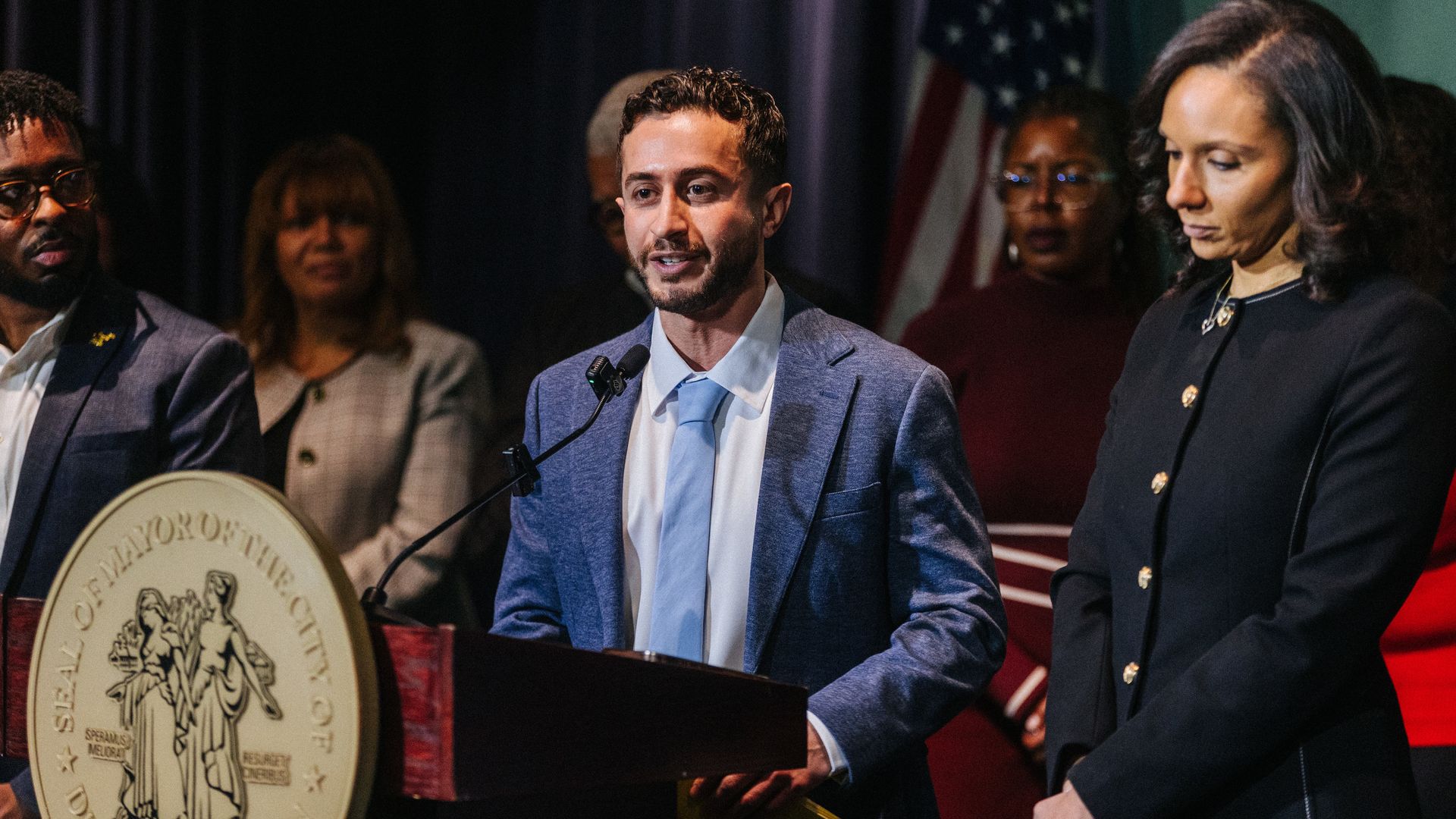 Man in a blue suit and light blue tie speaking at a podium with the Seal of Mayor of Detroit. He's next to Mayor Mary Sheffield
