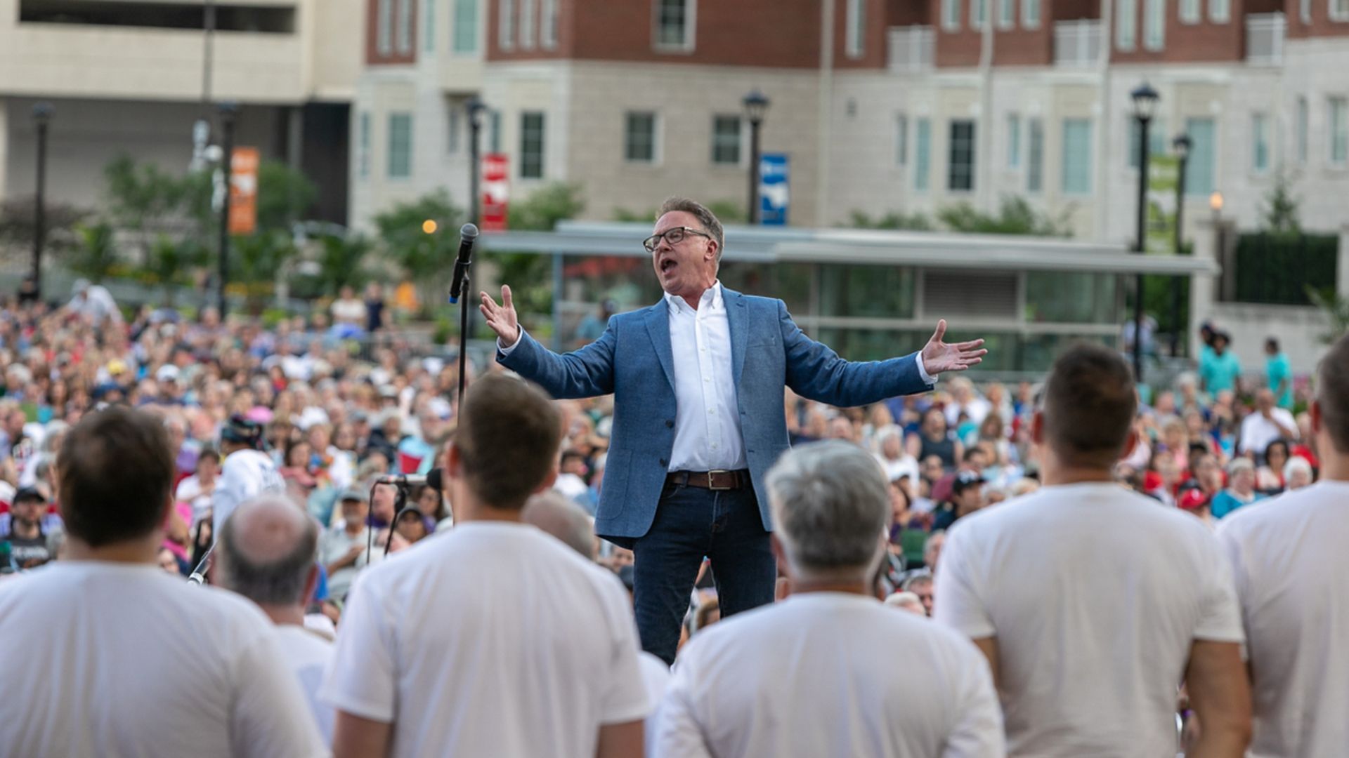 David Brown passionately directs the Harmony Project choir on an outdoor stage with microphone, with several singers in white shirts seen from behind.