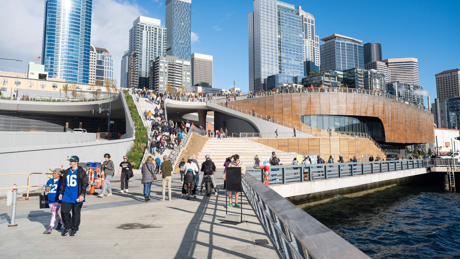 People walk by the Overlook Walk and the Seattle Aquarium's new Ocean Pavilion on the Seattle waterfront, with the Seattle skyline in the background.