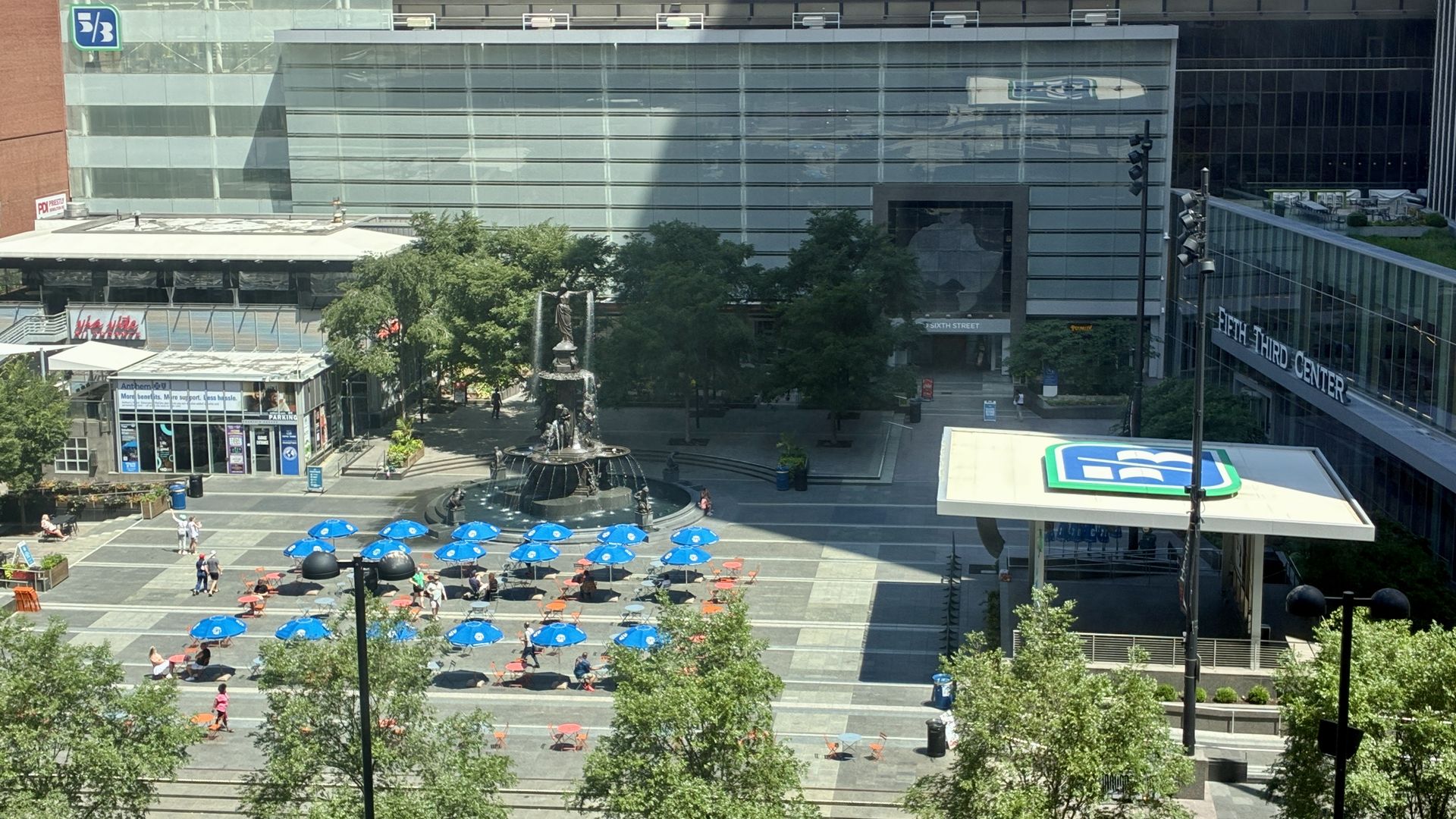 Sunny urban plaza, Fountain Square, with tall multi-tier Tyler Davidson Fountain, green trees, and rows of blue and orange umbrella tables. Modern glass buildings, Sixth Street entrance, and The Third Center signage visible.