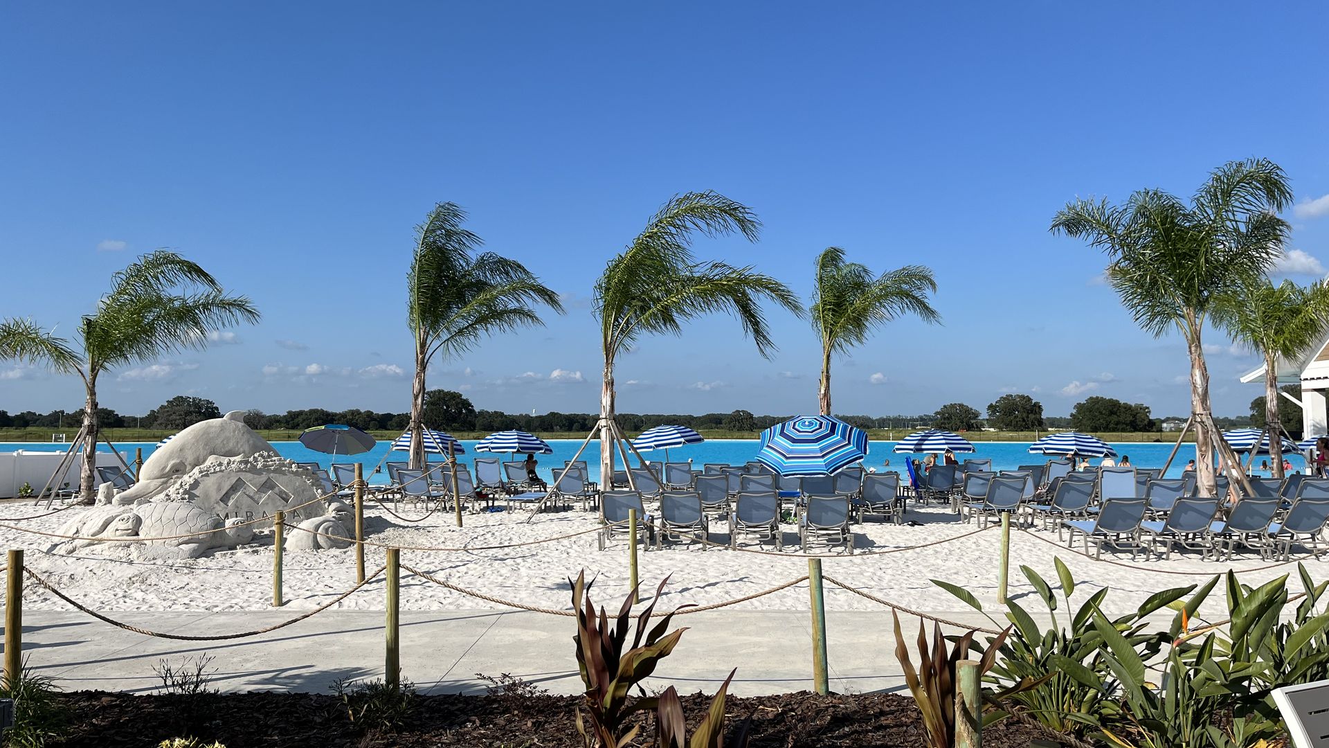A sandy shore with palm trees, lounge chairs, umbrellas and a sand sculpture. Beyond the shore is turquoise water.