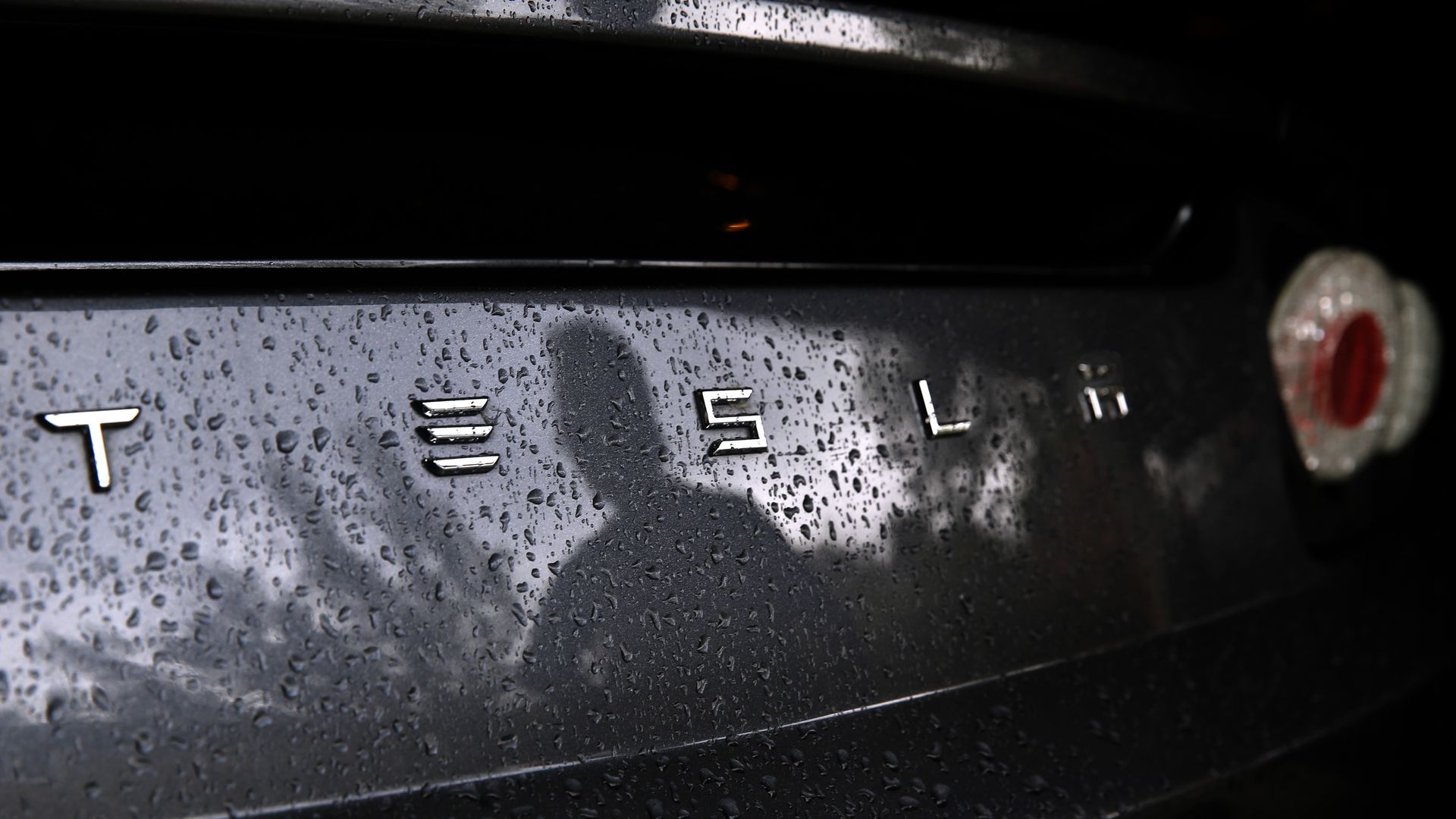 Close-up of a dark Tesla rear, covered in raindrops; silver TESLA lettering along the trunk and a circular red taillight visible on the right.