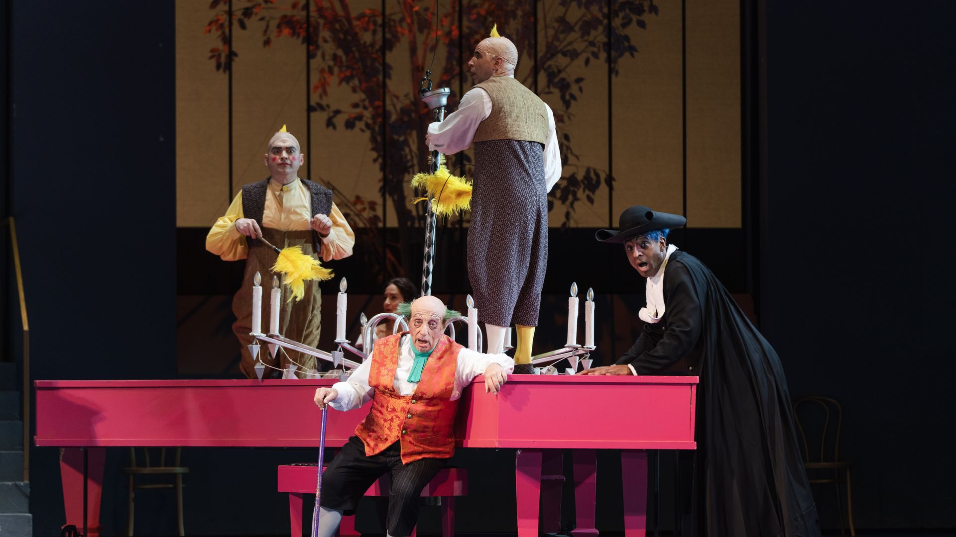 Clowns perform on a bright pink stage; a bald performer with a yellow feather Mohawk holds a tall staff while two others in colorful costumes watch, with a painted tree backdrop.