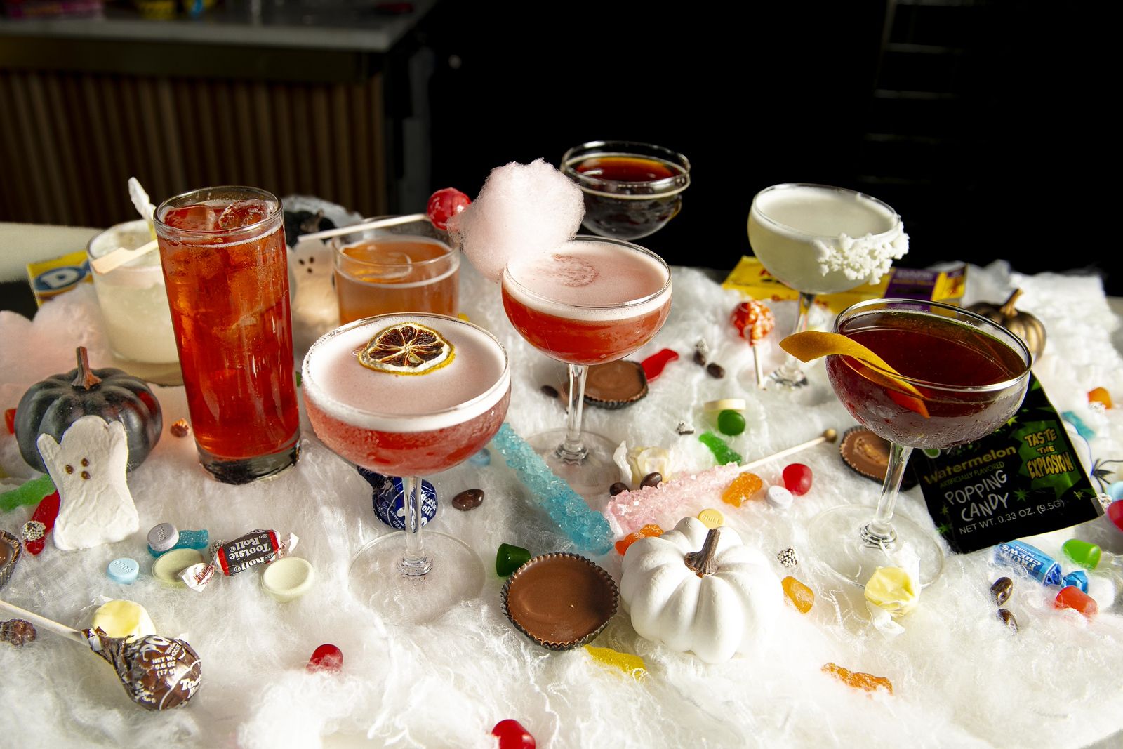 A Halloween-themed display of colorful cocktails surrounded by candy, small white and black pumpkins, and decorative cotton resembling spider webs on a table.