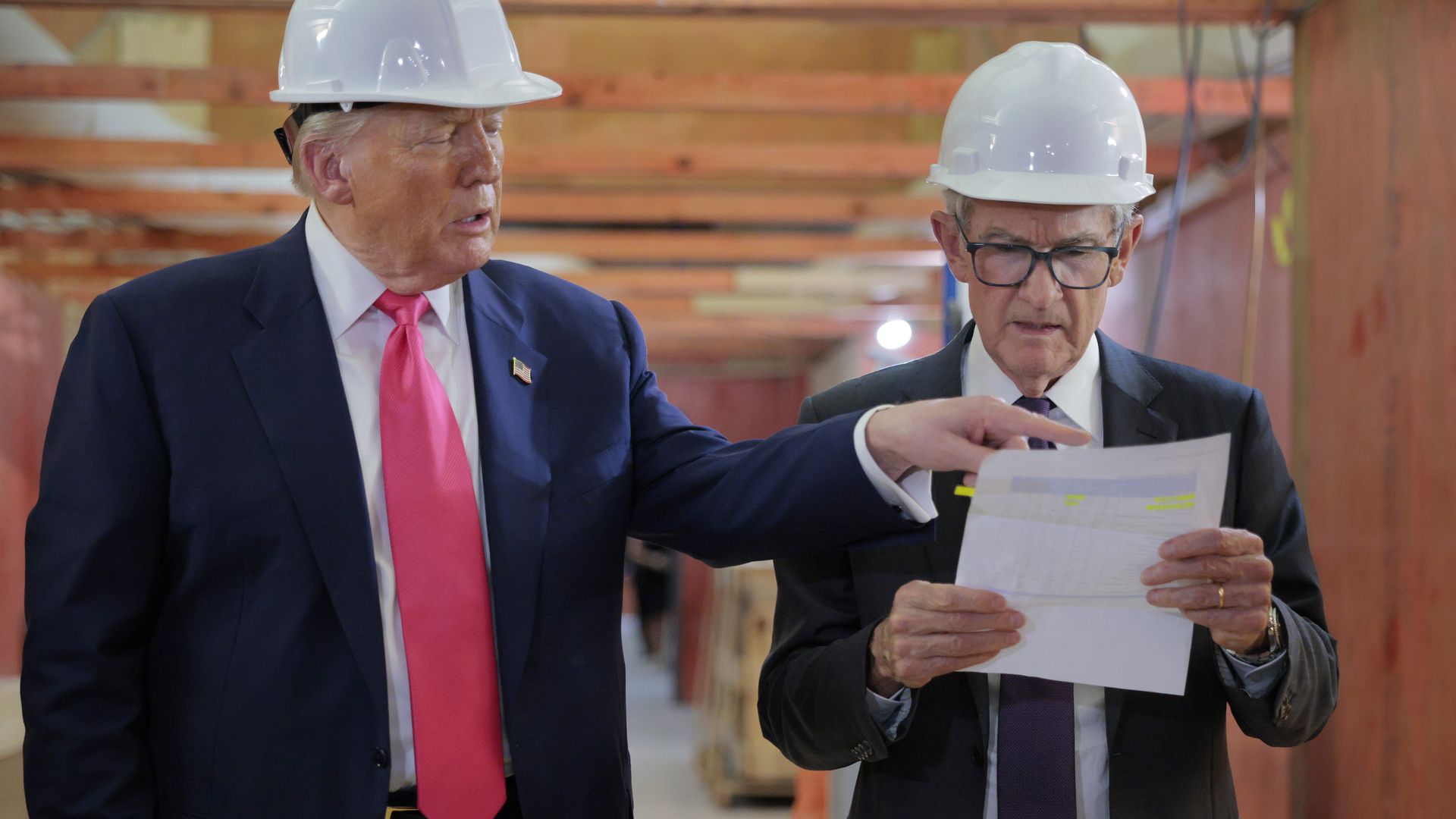 President Trump and Fed chair Jerome Powell, in suits and white hard hats inside a building under construction, Trump pointing at a document Powell is reading.