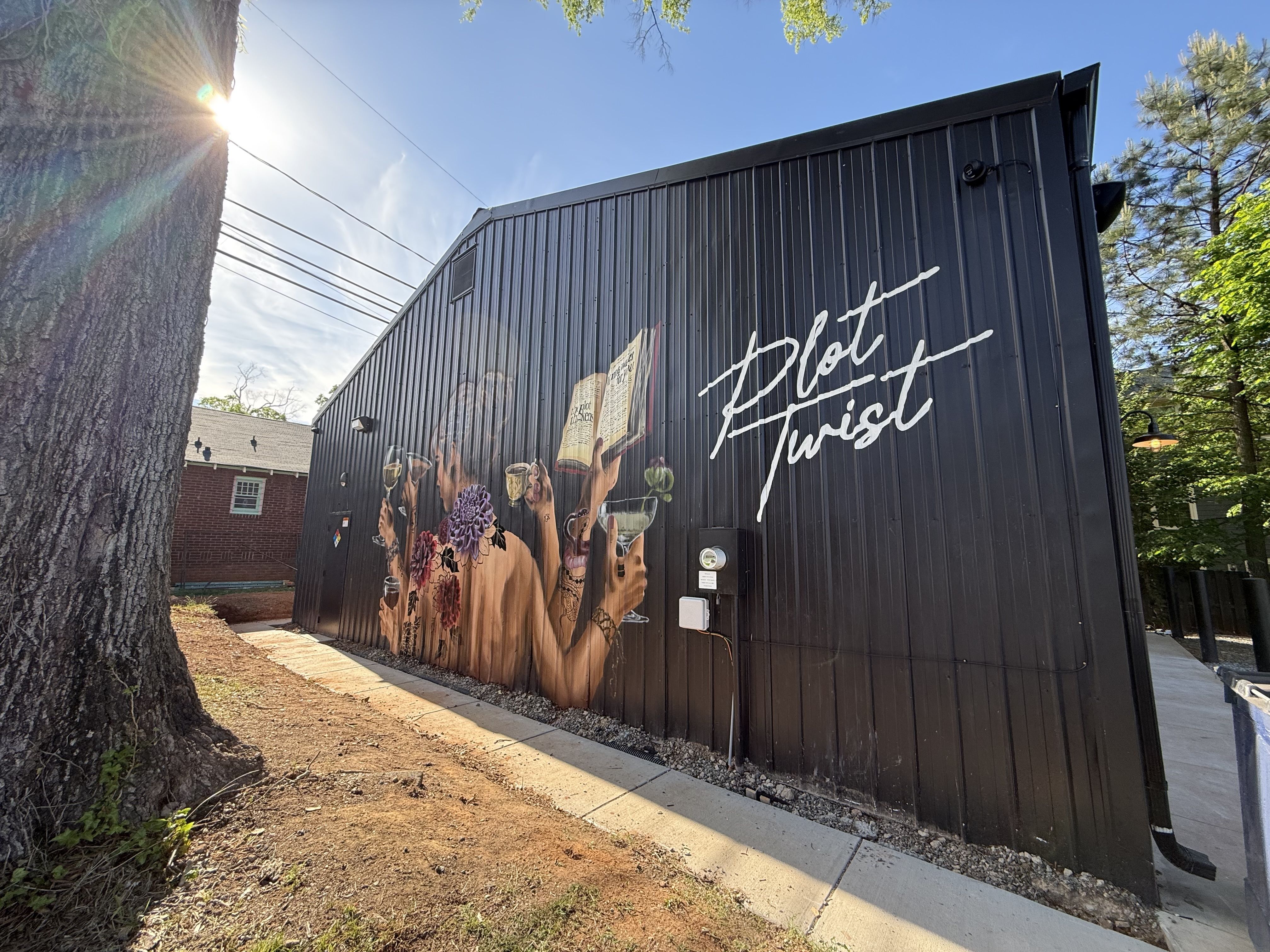 Black metal wall mural showing people raising glasses with flowers on their bodies and an open book; white cursive "Hot Twist" text on the right; sunny day, trees and a large tree trunk to the left.