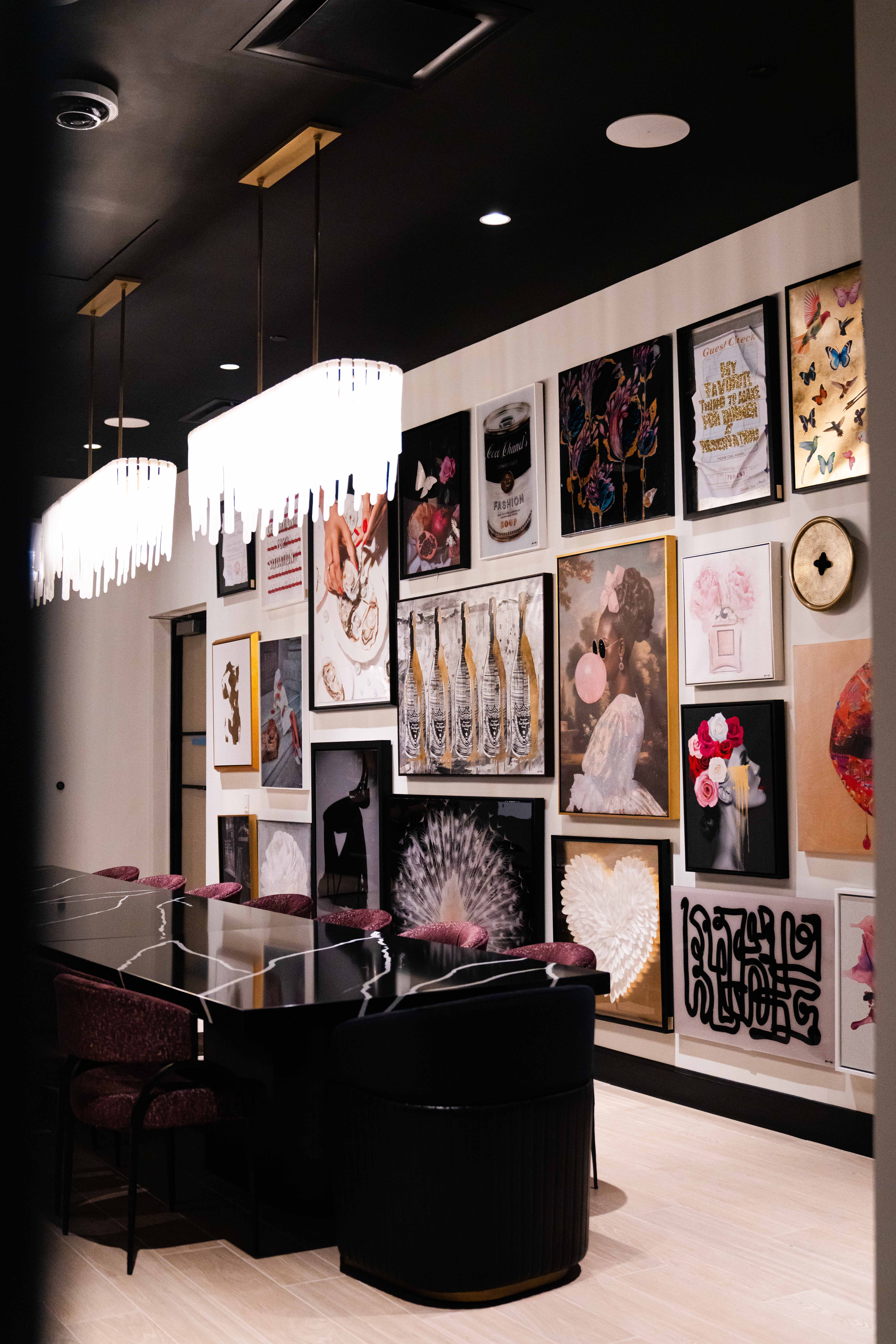 Modern dining room with long black marble table, purple chairs, two pendant lights, and a gallery wall featuring colorful artwork including flowers, butterflies, and abstract designs.