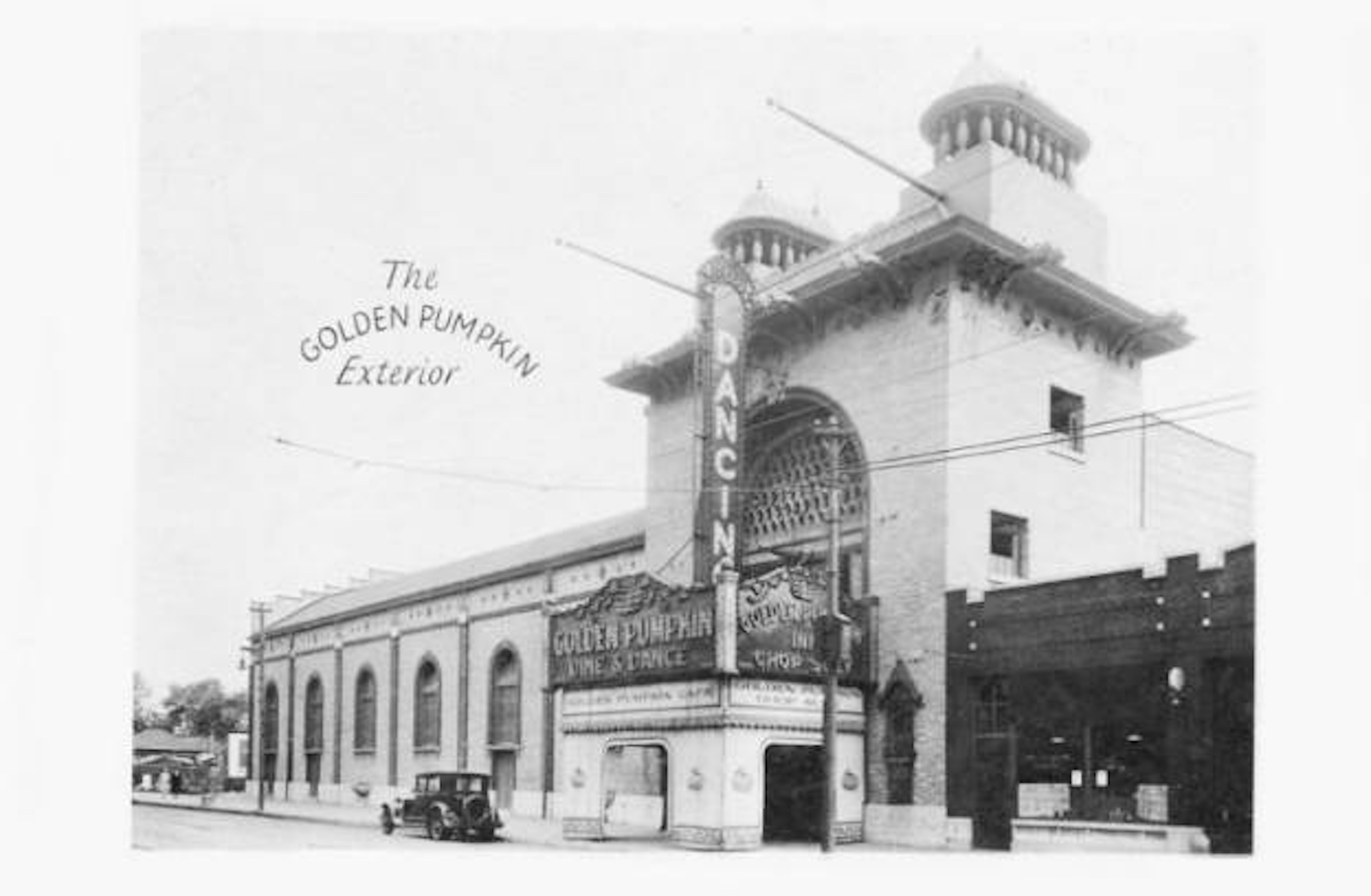 Black-and-white street scene of a vintage theater with arched windows and ornate cornice. Sign reads "The Golden Pumpkin Exterior" and vertical "GOLDEN PUMPKIN" marquee; a car is parked outside.