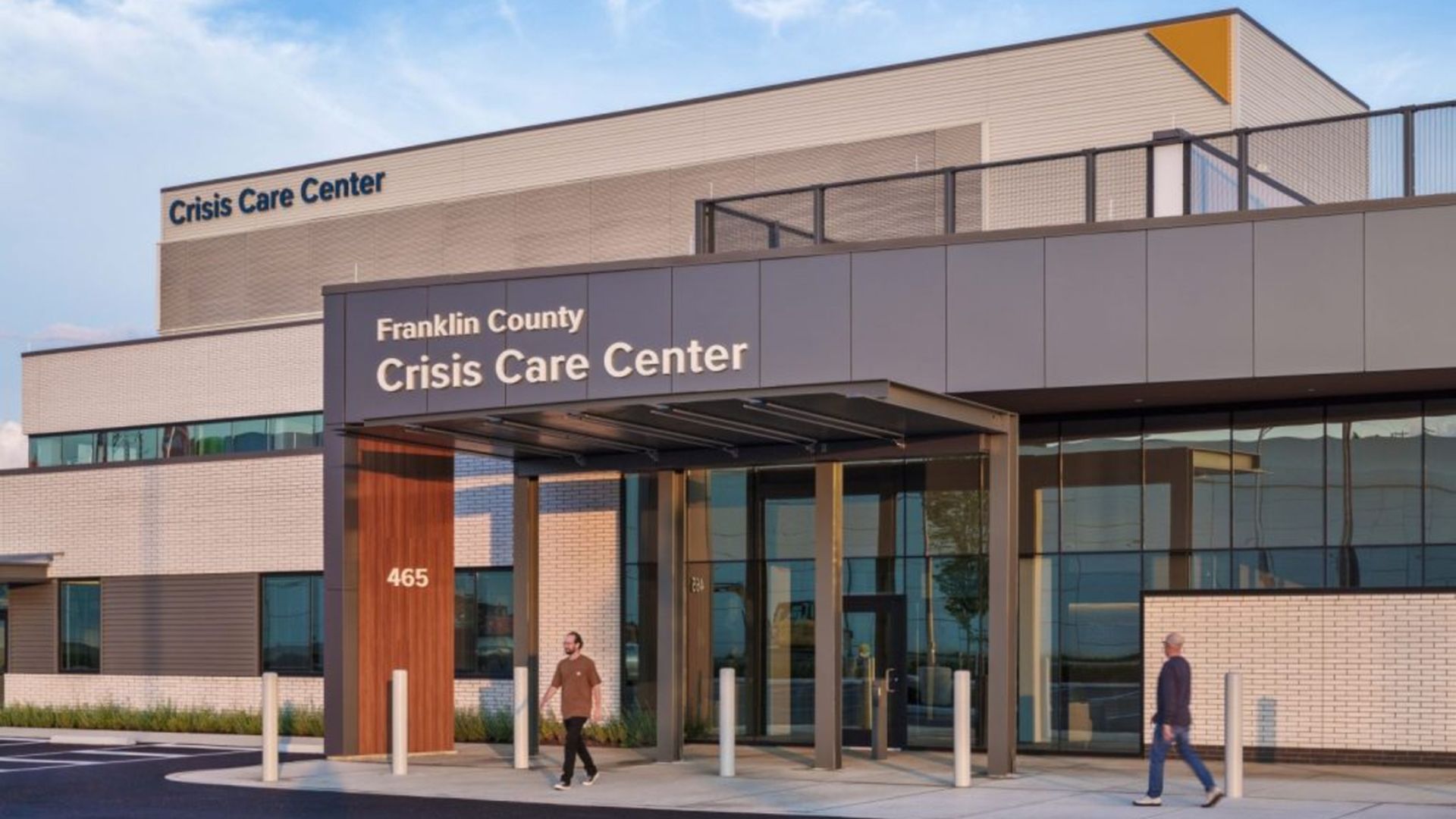 Exterior of a modern crisis care center with a glass entrance and two pedestrians walking by; signs read "Franklin County Crisis Care Center" and "Crisis Care Center" against a blue sky.
