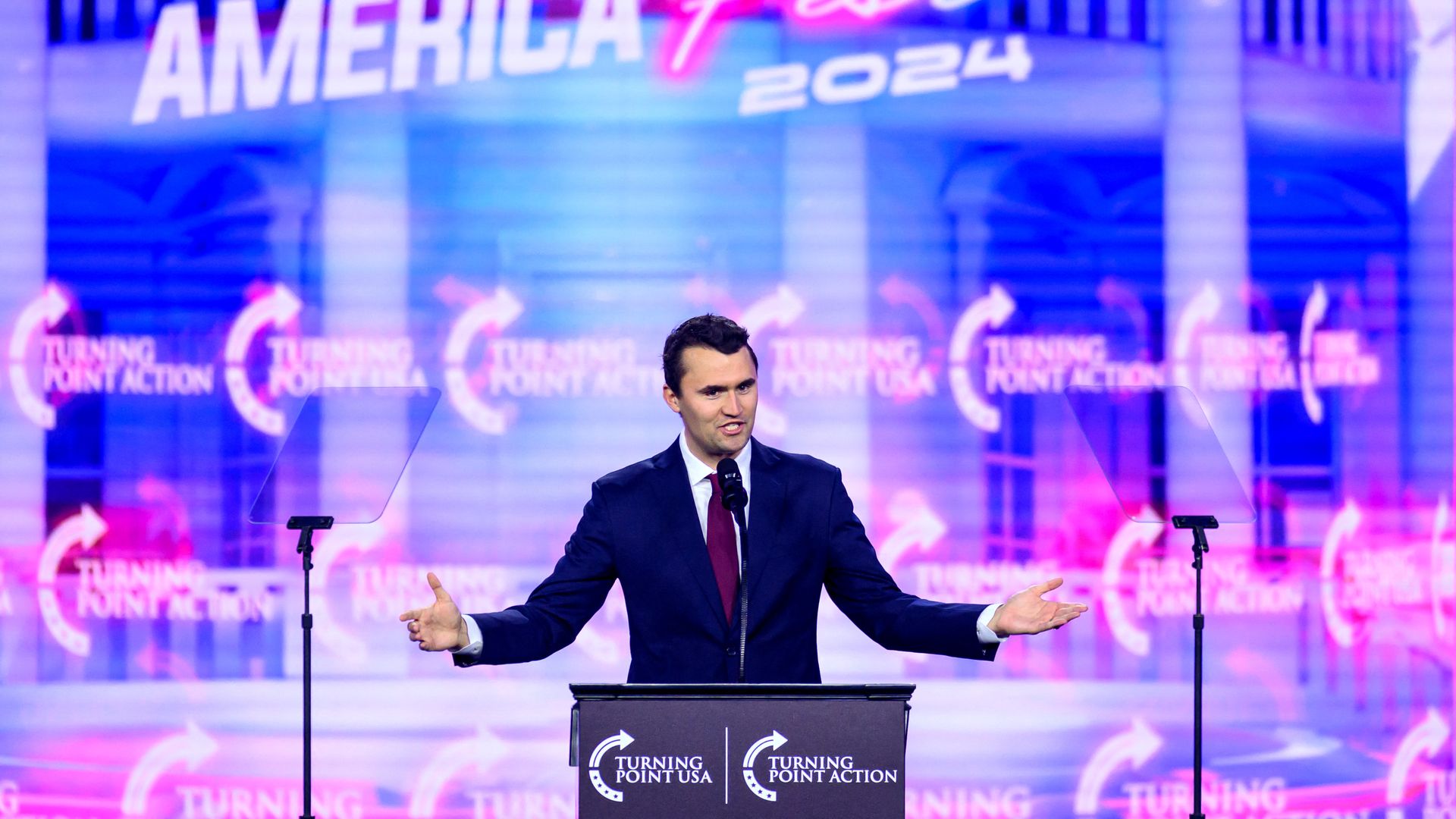 Charlie Kirk, wearing a blue suit and standing in front of a neon blue, white and red backdrop behind a podium and two glass teleprompters.