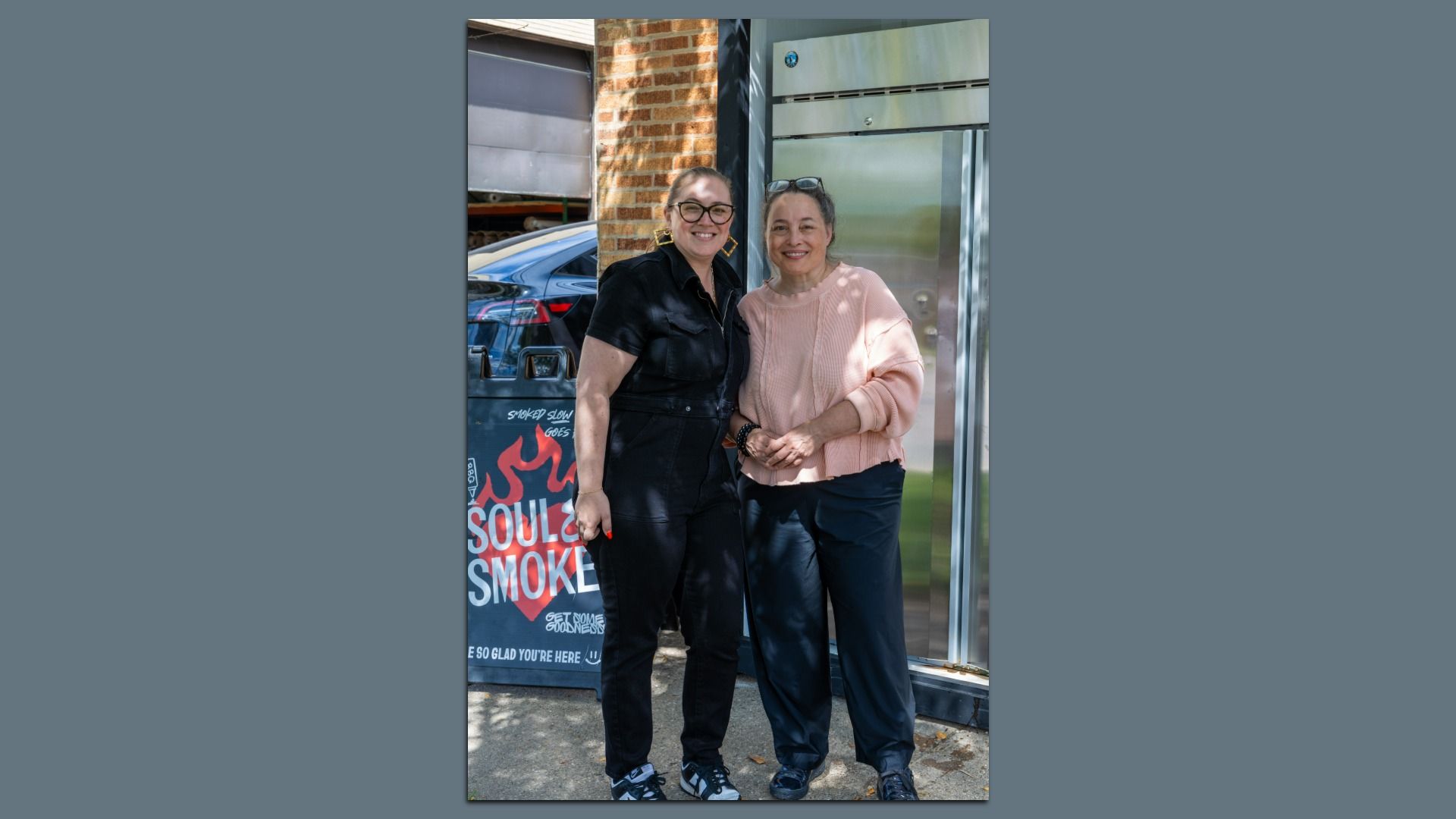 Two women standing outside near a brick wall and metal door. One wears a black outfit with glasses, the other a pink sweater and dark pants. A sign behind reads "Soul & Smoke."