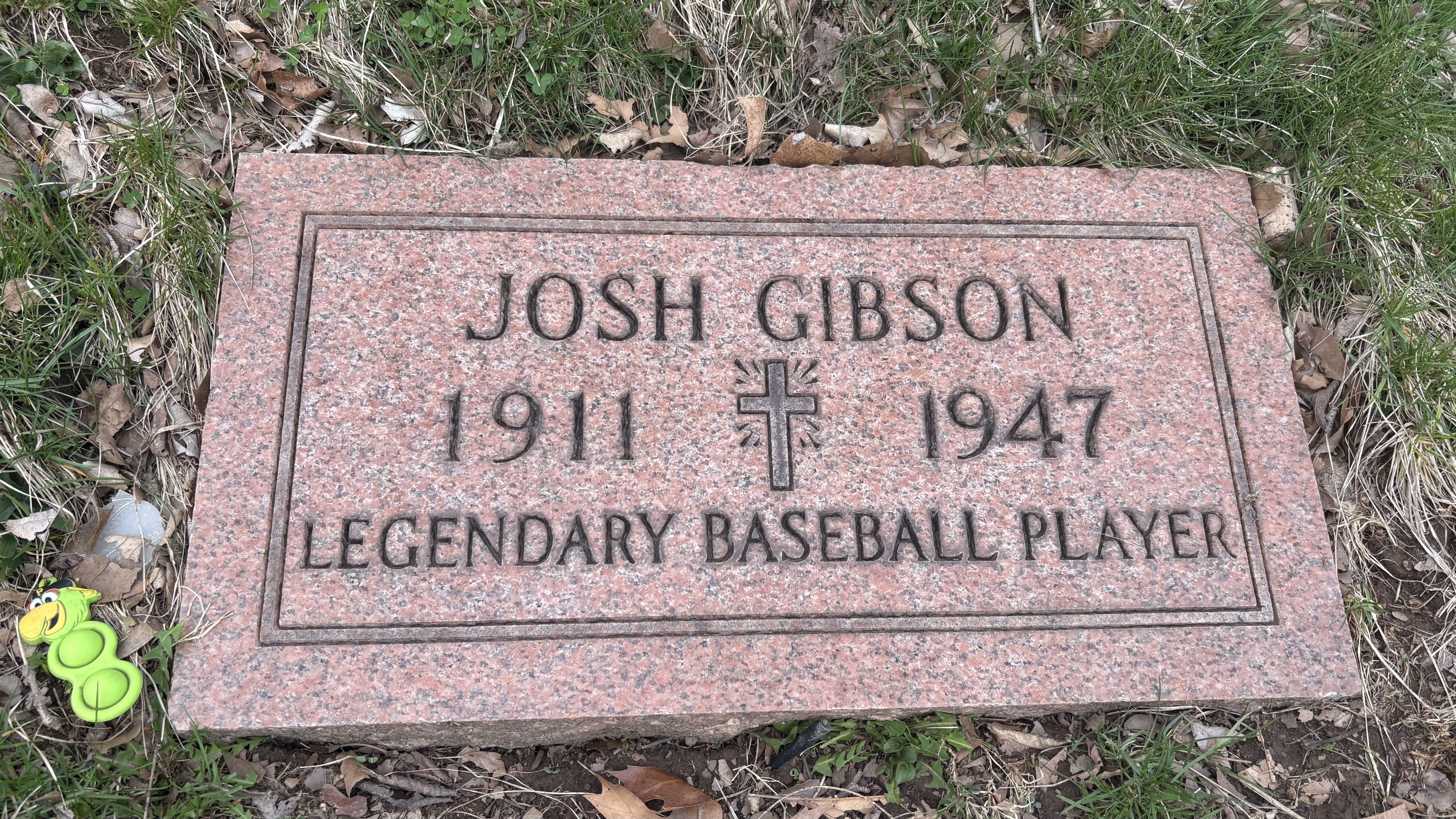 Pink granite gravestone of Josh Gibson (1911-1947), inscribed "Legendary Baseball Player," with a decorative cross between the years, surrounded by grass and leaves.