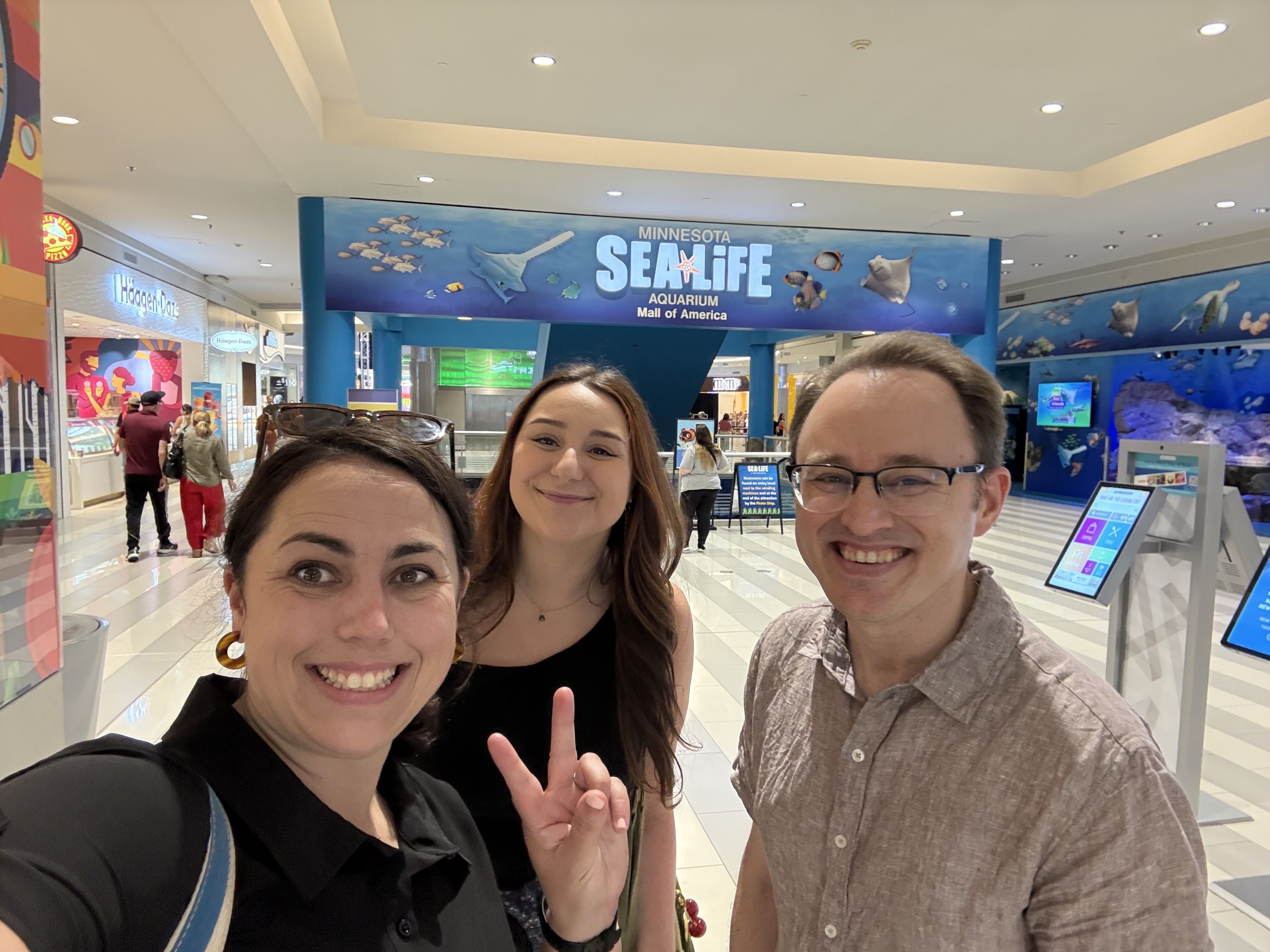 Three people take a selfie in a mall beside a large blue Minnesota Sea Life Aquarium sign; Haagen-Dazs storefront visible on the left.