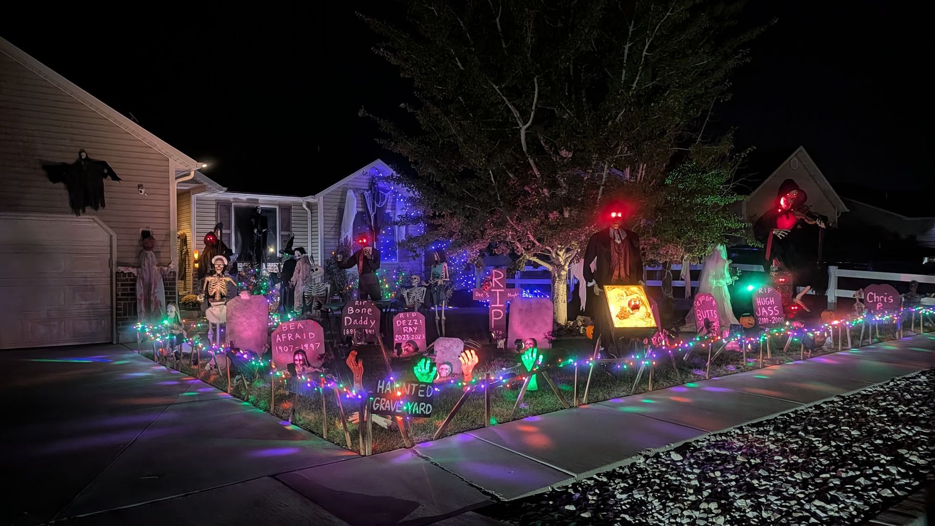 Night image of a suburban front yard Halloween display: skeletons, tombstones, ghosts, and colorful lights along a fence; eerie red-eyed figures stand among the decorations near a tree and house.
