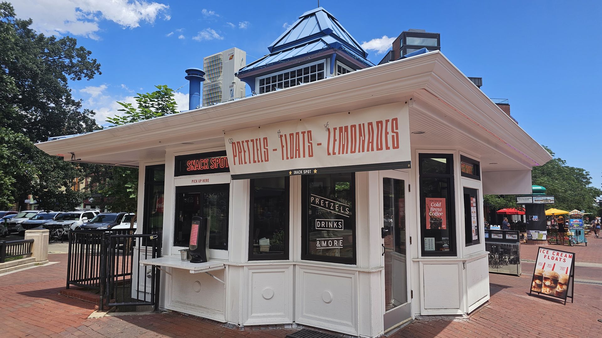 White corner snack stand with a large banner advertising pretzels, floats, and lemonades in red letters. Windows display snacks and drinks; blue sky, green trees, and parked cars nearby.
