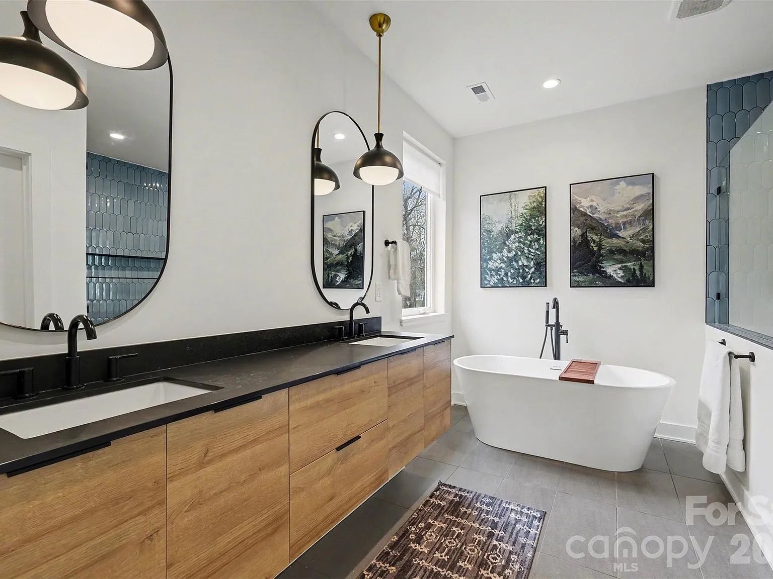Modern bathroom with long wooden vanity, black countertop, two oval mirrors, matte black fixtures, white freestanding tub with wooden tray, blue tiled shower, and nature artwork on walls.
