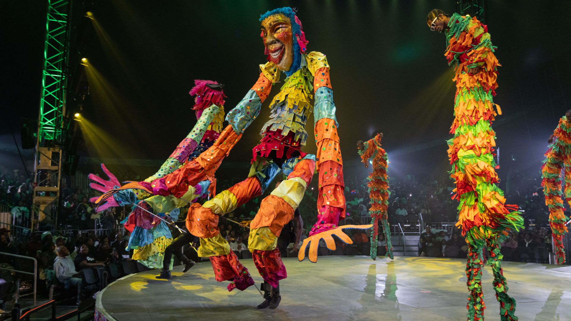 Vibrant stage show with oversized, multicolored puppets and dancers in fringed costumes. Central figure wears a golden mask with blue hair and a big smile, under green lights as the audience watches.