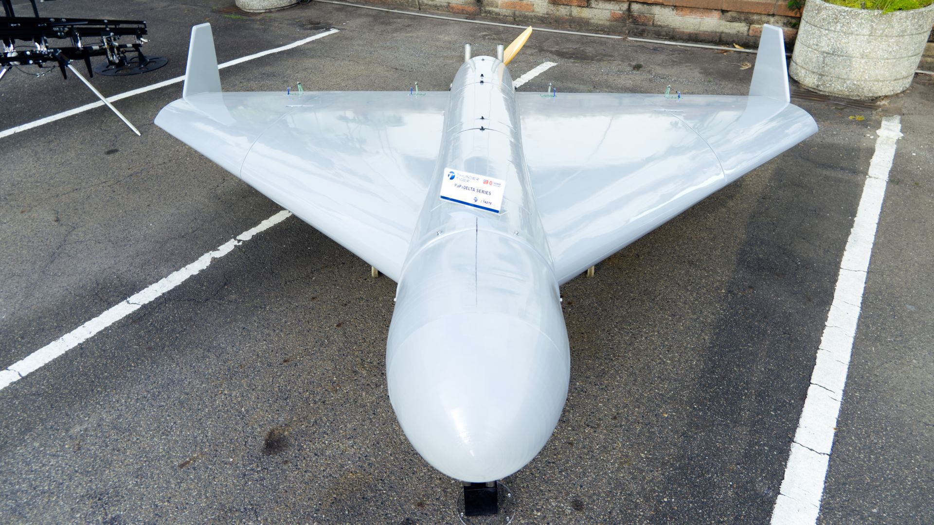 Top-down view of a large silver model airplane parked on an asphalt lot. It has a long fuselage, wide wings, twin vertical tails, and a yellow rear propeller; a label hangs from the fuselage.