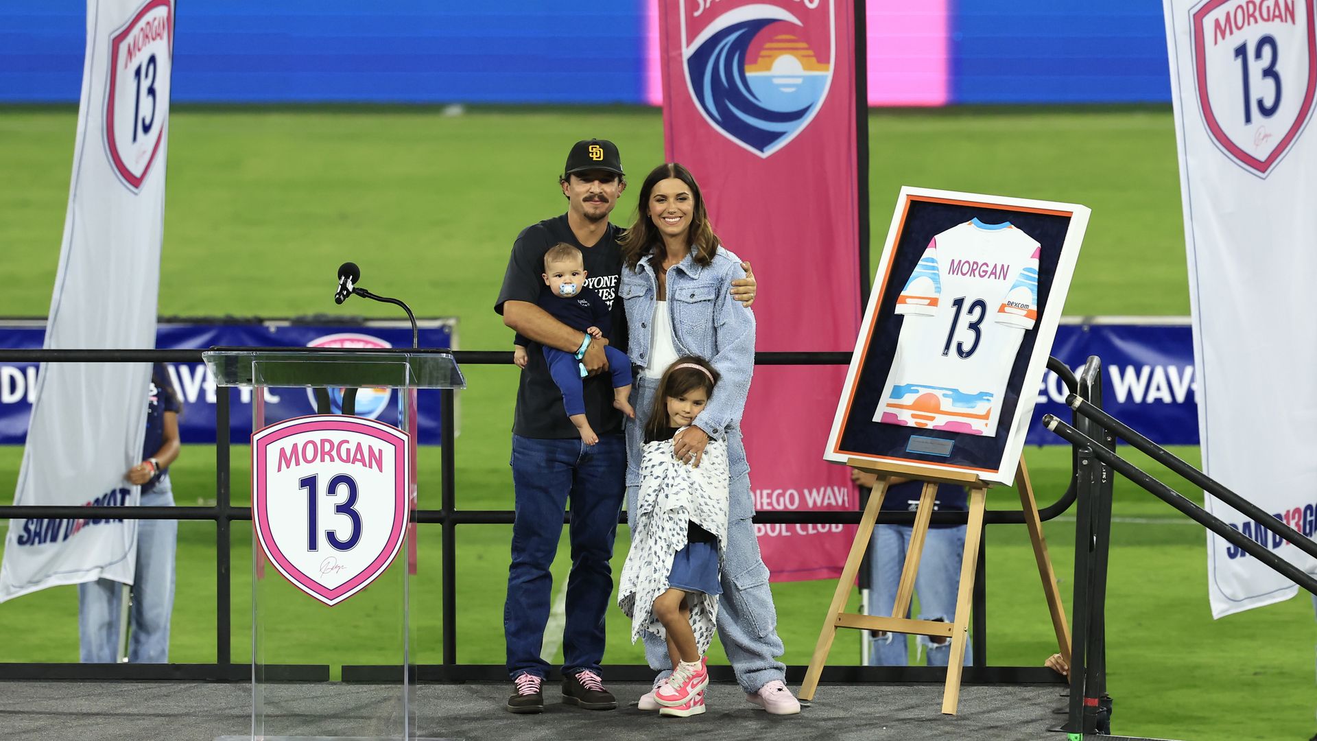 Alex Morgan stands on stage with her husband, Servando Carrasco, and children, her framed Number 13 jersey on an easel and San Diego Wave banners. 