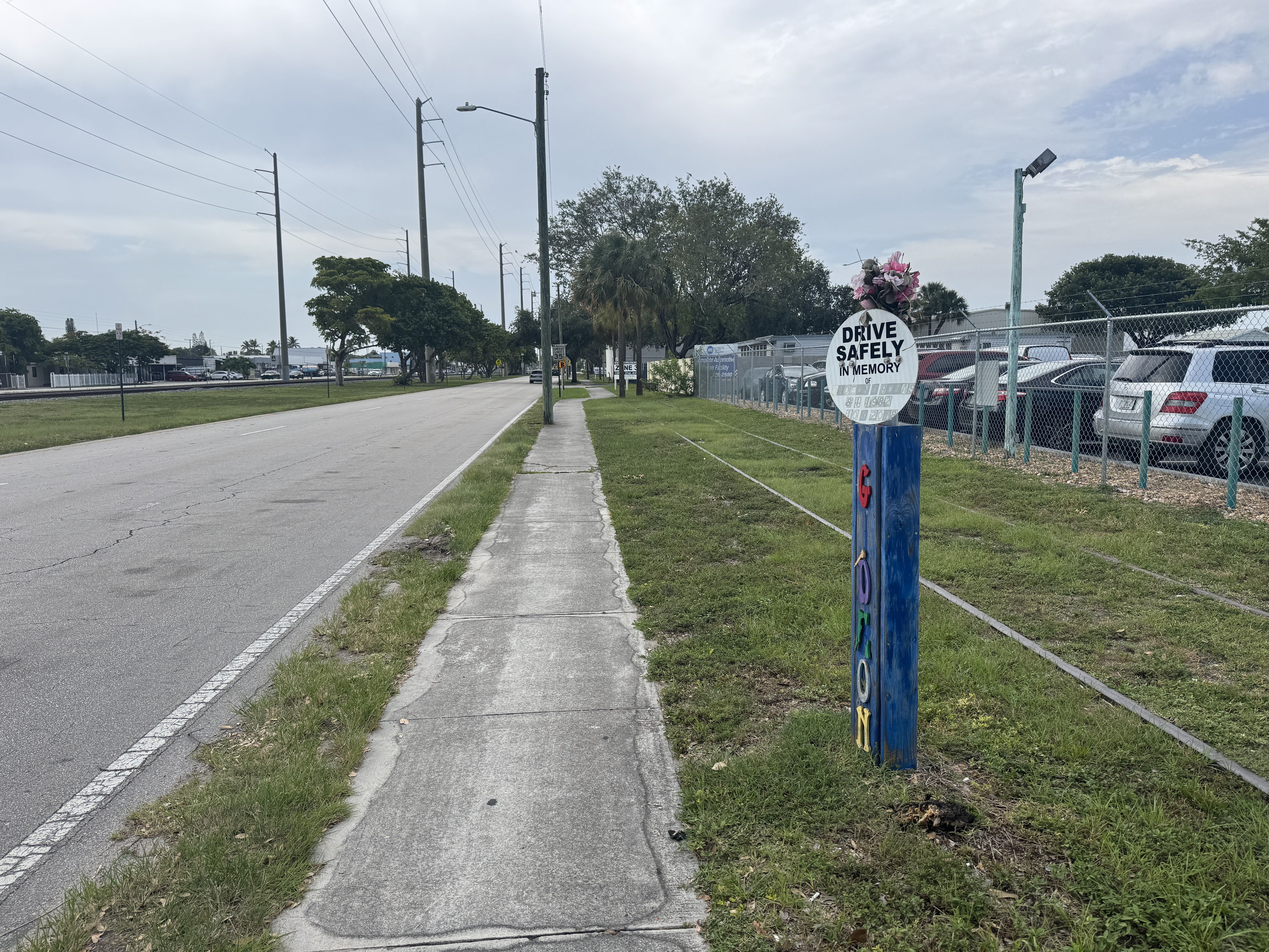 Wide street with cracked sidewalk, utility poles on the left, chain-link fence on the right, and a memorial sign reading "DRIVE SAFELY IN MEMORY OF" topped with pink flowers; cars behind the fence; overcast sky.