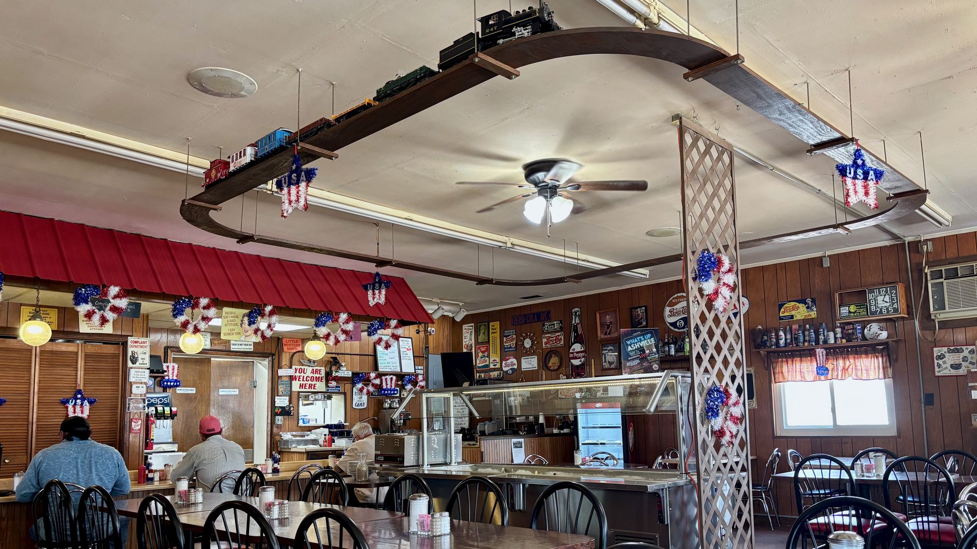 Retro diner interior with a suspended curved wooden model train track across the ceiling carrying miniature trains, red-white-blue decor, ceiling fan, wood-paneled walls, and diners at tables.