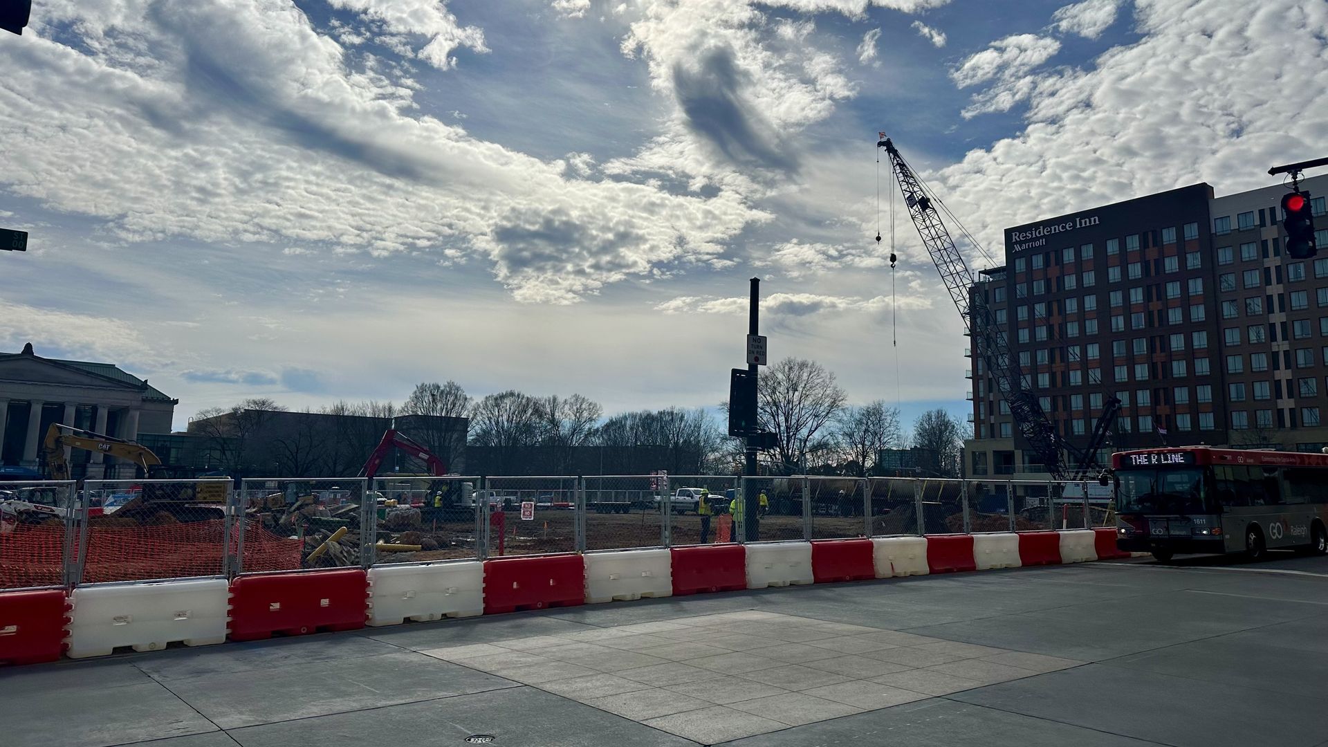 Construction site behind red and white barriers with workers, cranes, and machinery under a mostly cloudy sky, near a Residence Inn building and a public bus on the street.