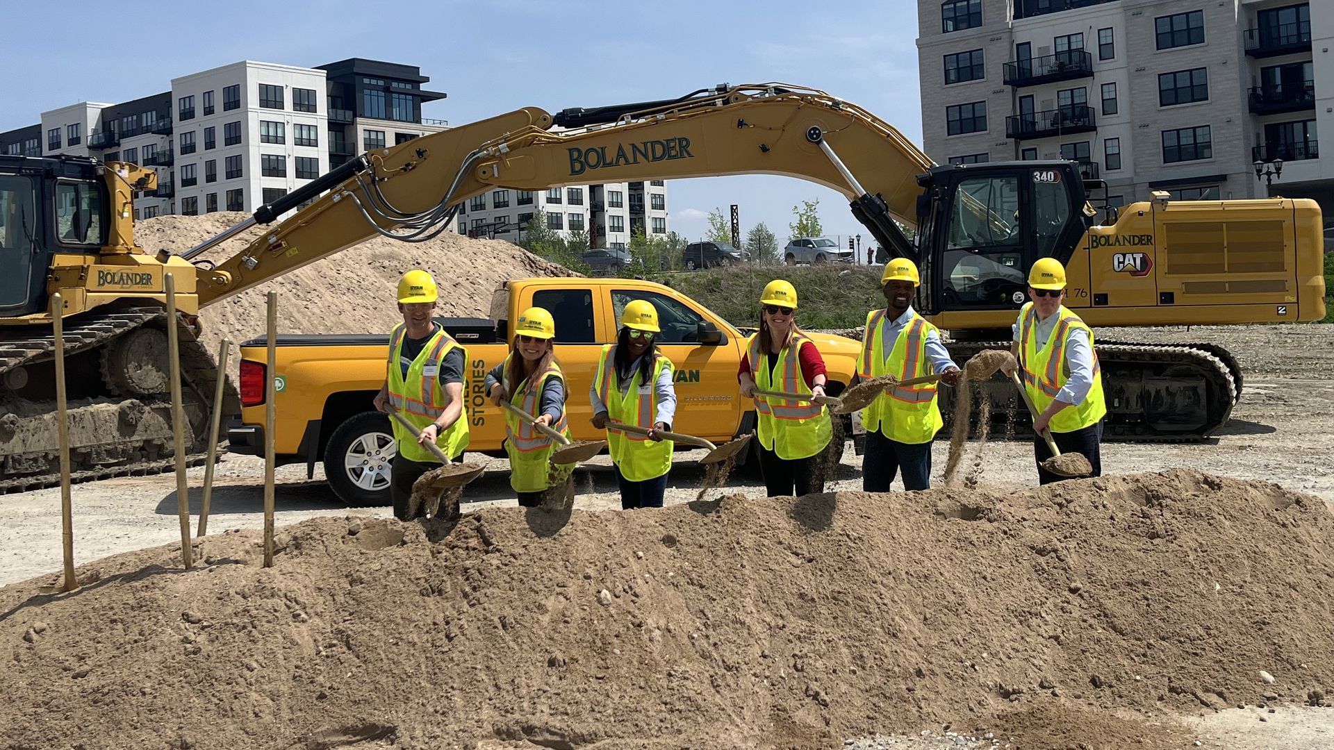 Officials hold shovels with dirt in front of a large excavator 