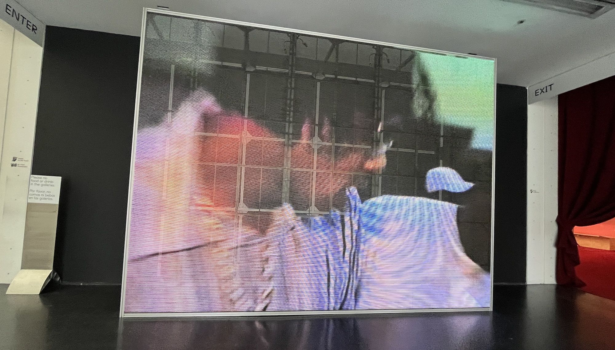 A large freestanding LED video screen in a gallery shows a blurry, gridlike scene of two people, one in blue with a cap, against black walls and a red curtain, near signs reading "ENTER" and "EXIT".