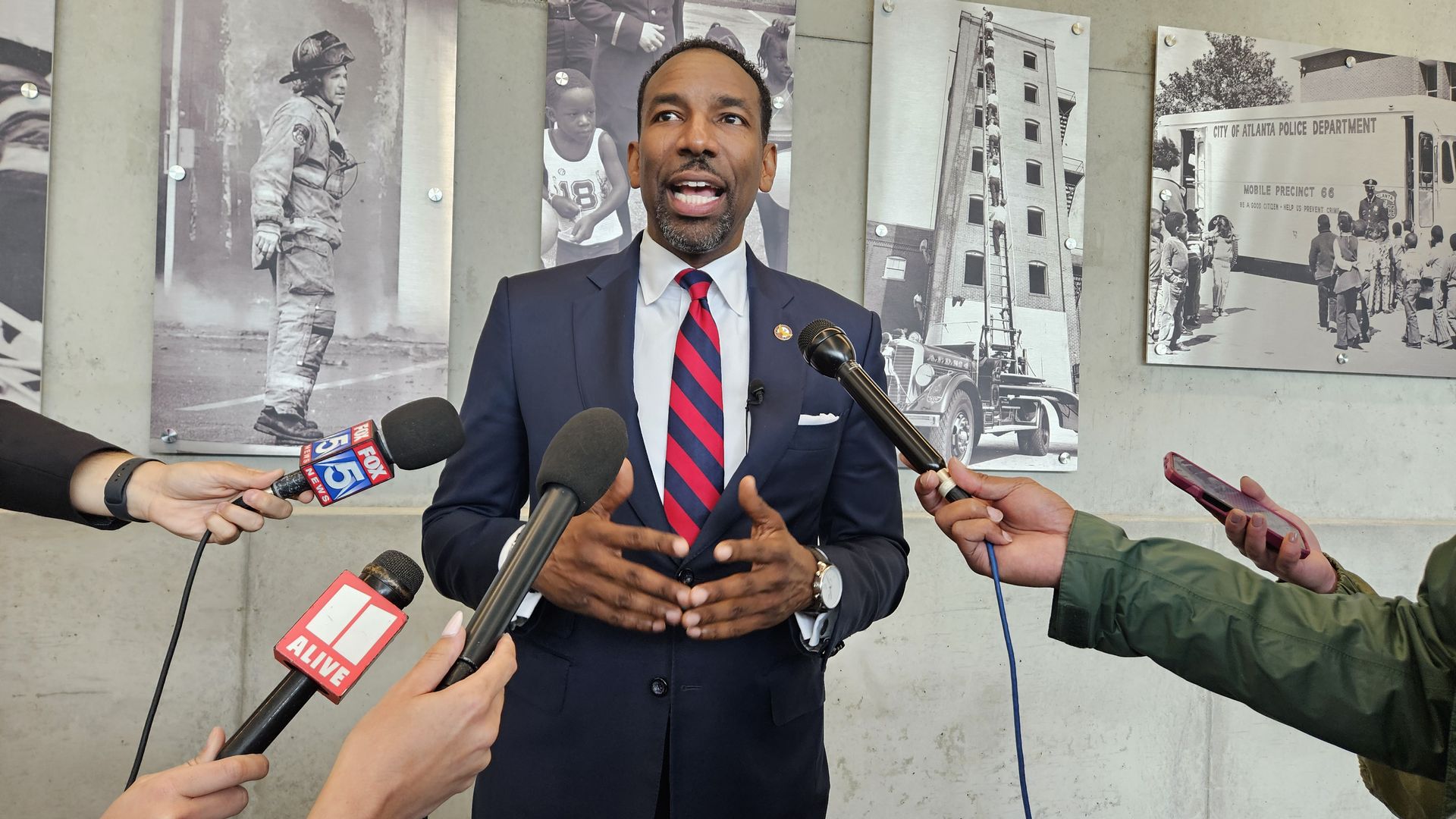 Man in a navy suit, white shirt, and red striped tie speaks to reporters holding microphones from FOX 5 News and 11 Alive, with black and white photos of firefighters and police behind him.