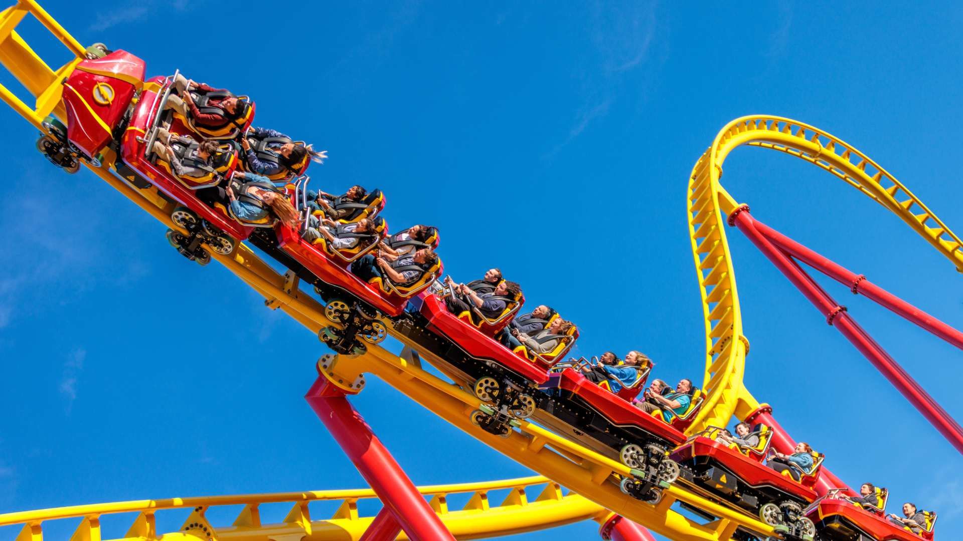 A red and yellow roller coaster train carries excited riders along a bright yellow track with a looping curve, set against a clear blue sky.