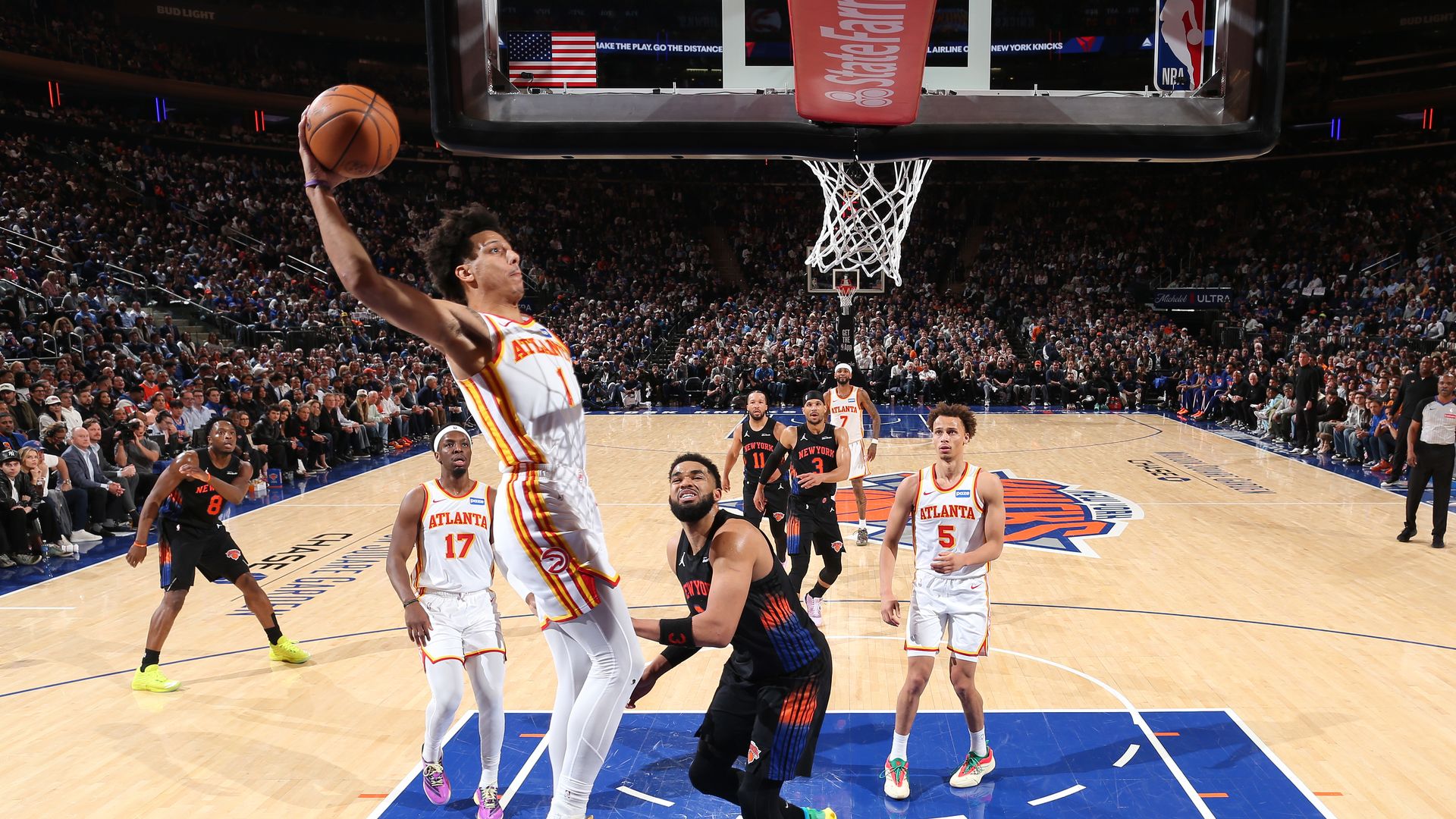 A basketball player wearing a white uniform attempts to slam dunk as a defender wearing a dark uniform looks on.