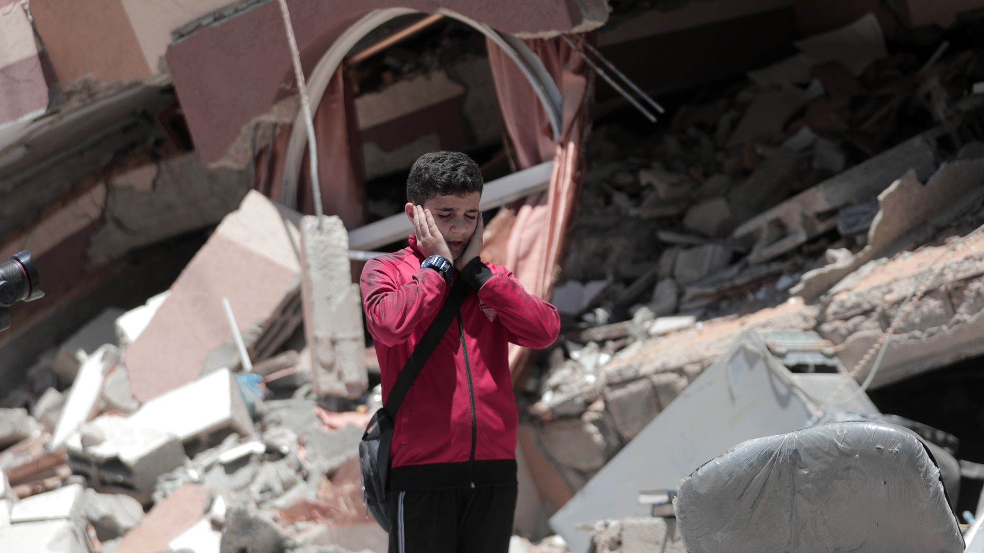Photo of a young Palestinian standing with their hands held over the ears amid rubble