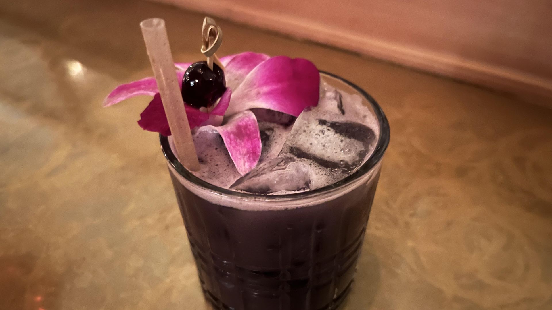 A dark purple cocktail in a glass with ice, garnished with a pink orchid flower and a dark cherry on a wooden pick, set on a reflective golden surface at Tokyo Cowboy.