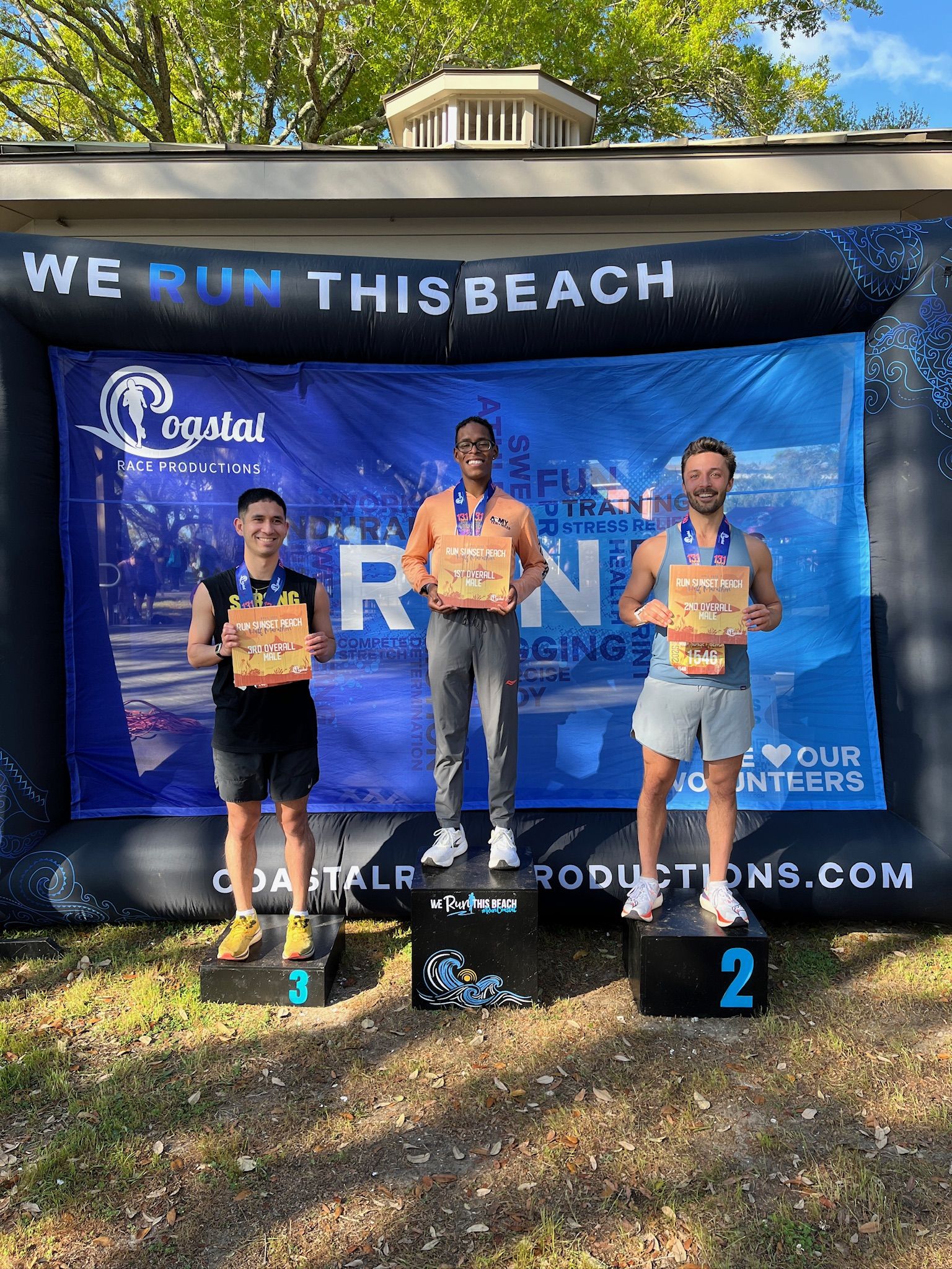 Three male runners stand on a podium outdoors under a "WE RUN THIS BEACH" arch, holding certificates. Center: orange shirt; 2nd: blue; 3rd: black. Blue Coastal Race Productions backdrop behind.