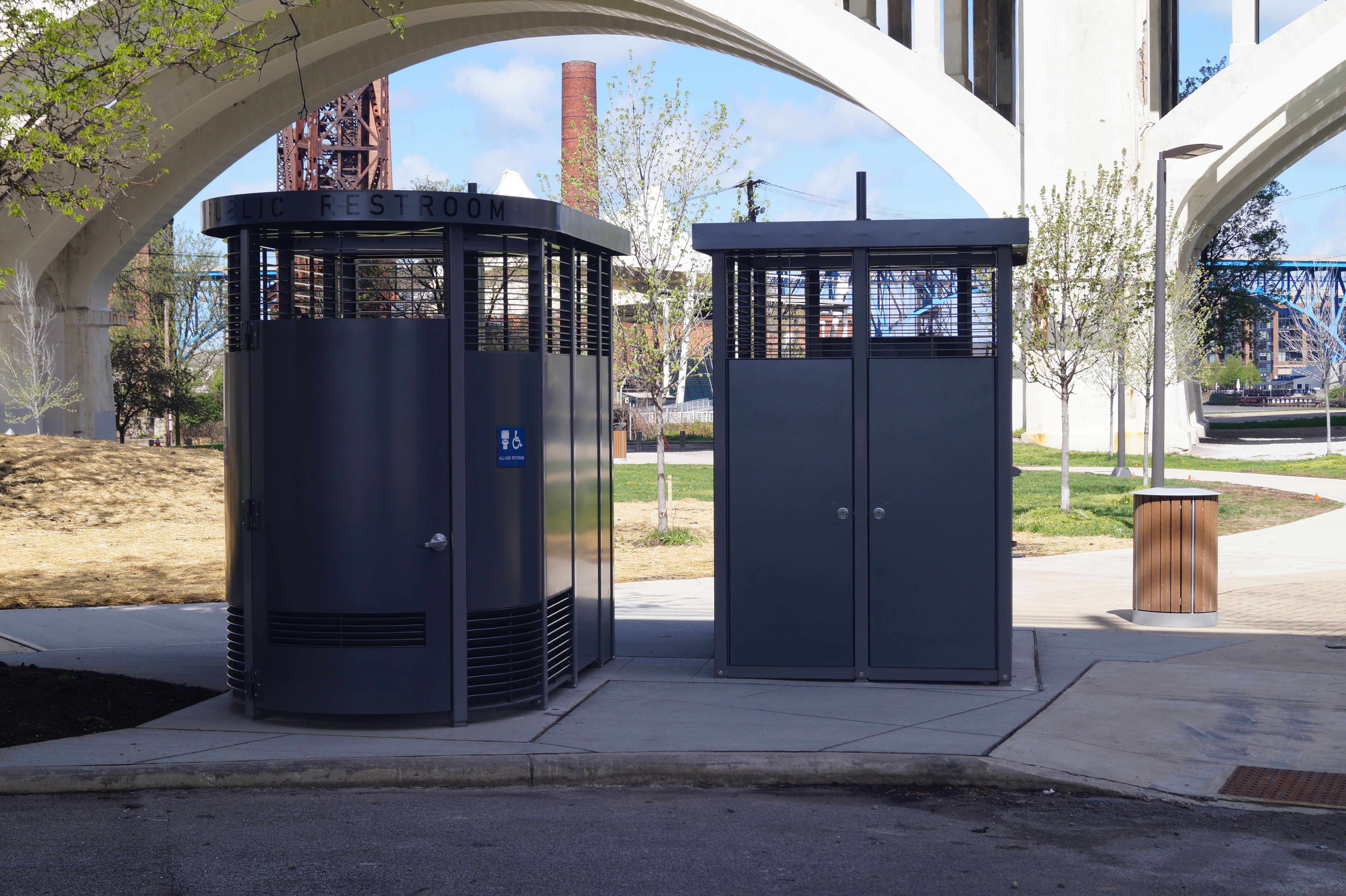Two dark gray public restrooms with slatted walls sit beneath a large white concrete arch. A blue wheelchair sign is on the left unit; trees and distant industrial structures frame the background.