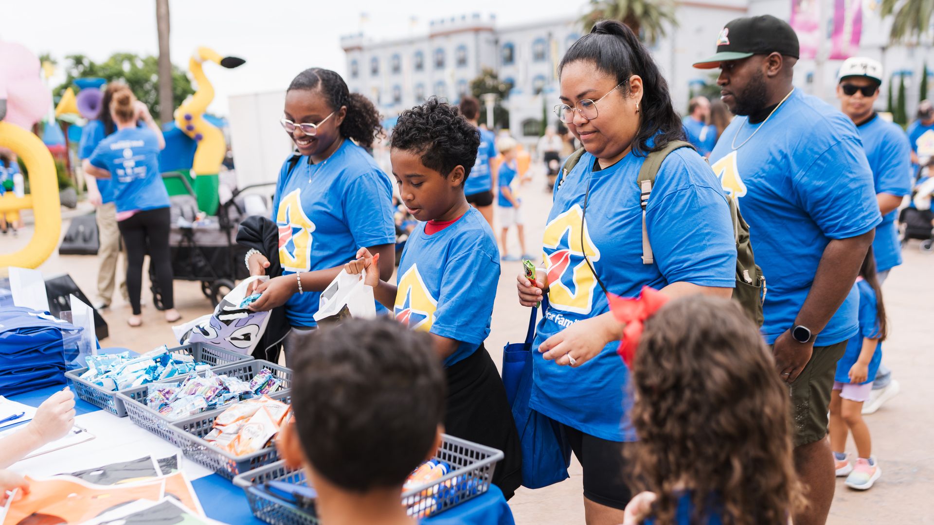 Volunteers in blue shirts hand out snacks at an outdoor event. A woman with glasses and a child stand by snack baskets, with colorful inflatables and palm trees in the background.