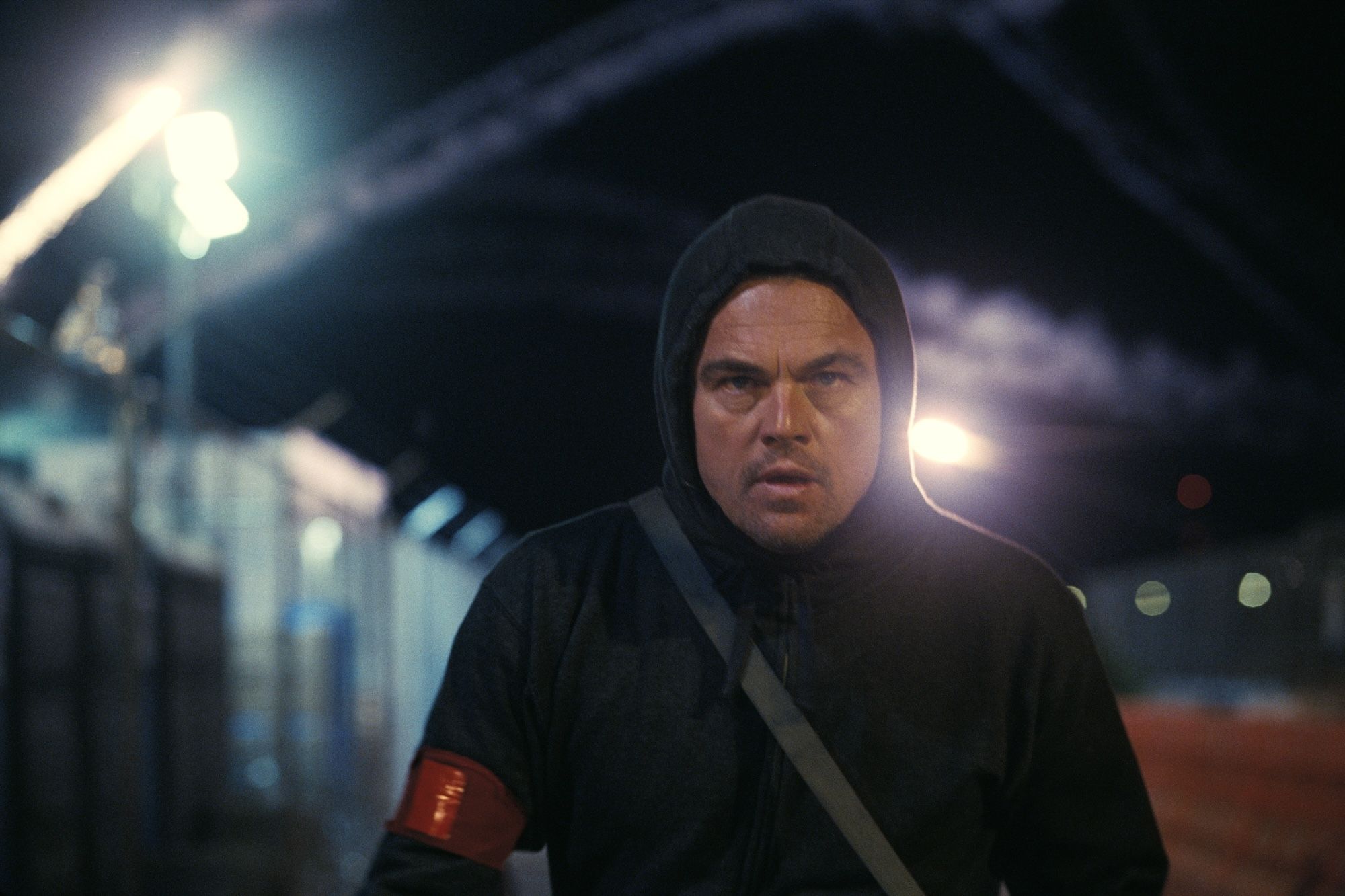 Man with determined expression wearing a black hoodie and red armband, walking at night near a fence with bright lights and smoke trails in the background.