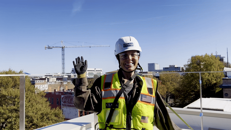 Smiling construction worker wearing a white hard hat and yellow safety vest with American flag patches, waving with a cityscape and crane in the background under clear blue sky.