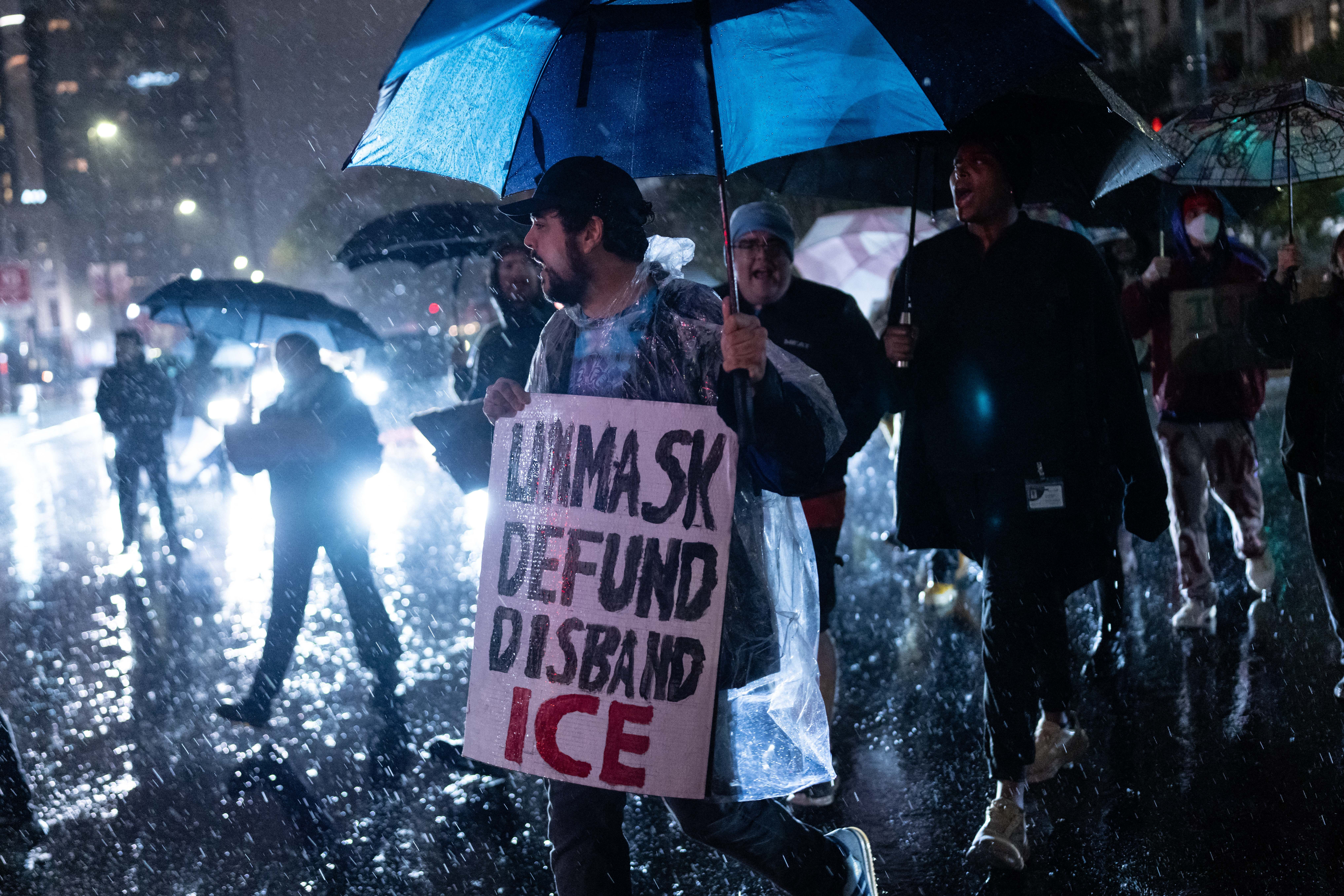 A small crowd is seen walking in the rain at night. One person, holding an umbrella, holds a sign that says "unmask, defund, disband ICE." 